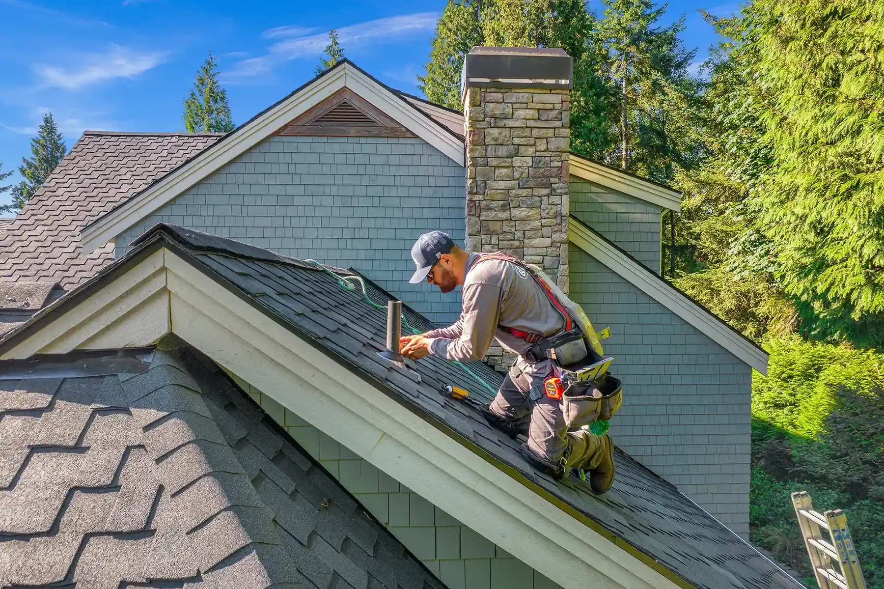 A worker inspecting or repairing a shingled roof of a house surrounded by trees under a clear blue sky.