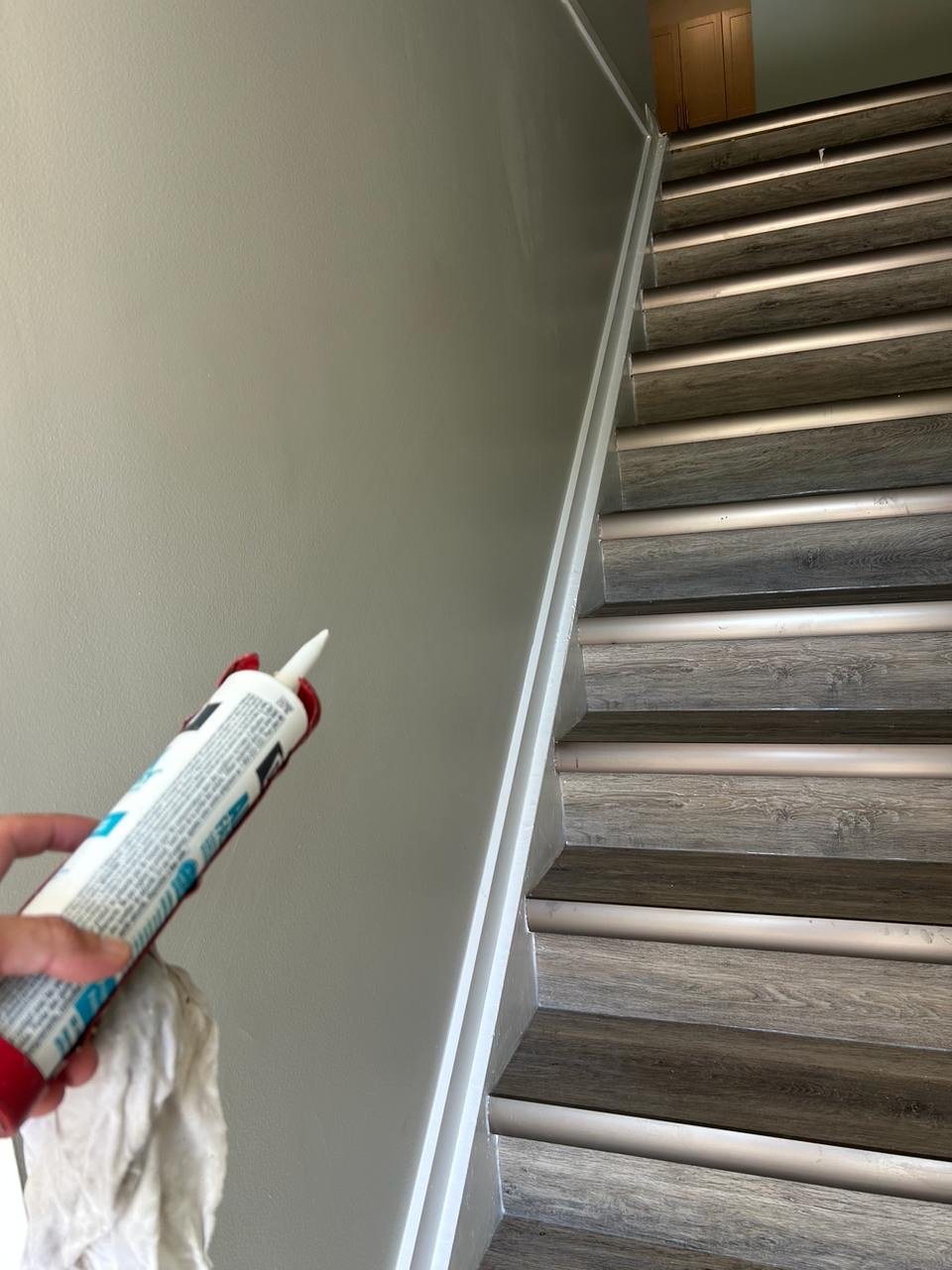 A person holding a caulking gun and a cloth near a staircase with wood and white pipes.