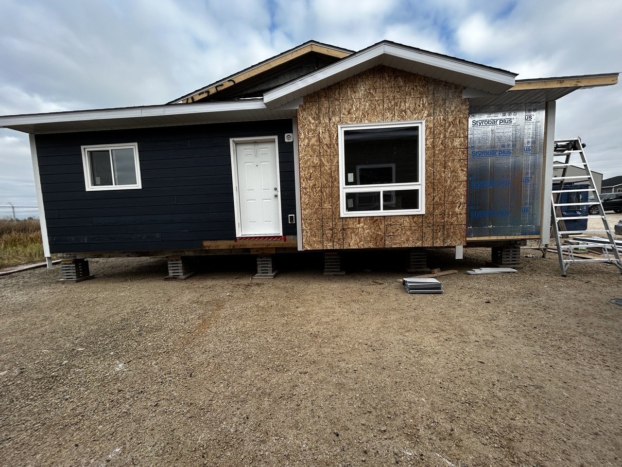 A house under construction with a black exterior on the left, some unpainted plywood on the right, and a window and door installed, all elevated on concrete blocks.