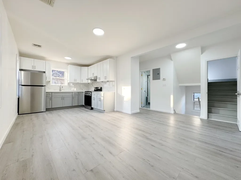 Empty, modern kitchen and living space with white cabinets, stainless steel refrigerator, stove, white walls, light wood flooring, and stairs leading to an upper level.