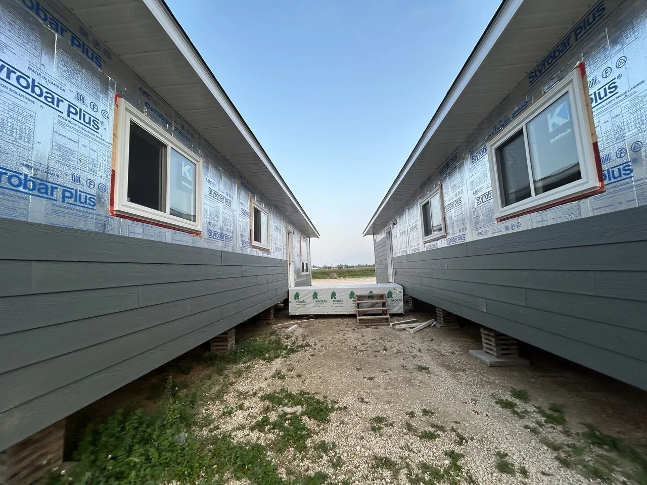 Two houses under construction, viewed from the ground level, with unfinished bases and windows installed, facing each other, and an open sky above.