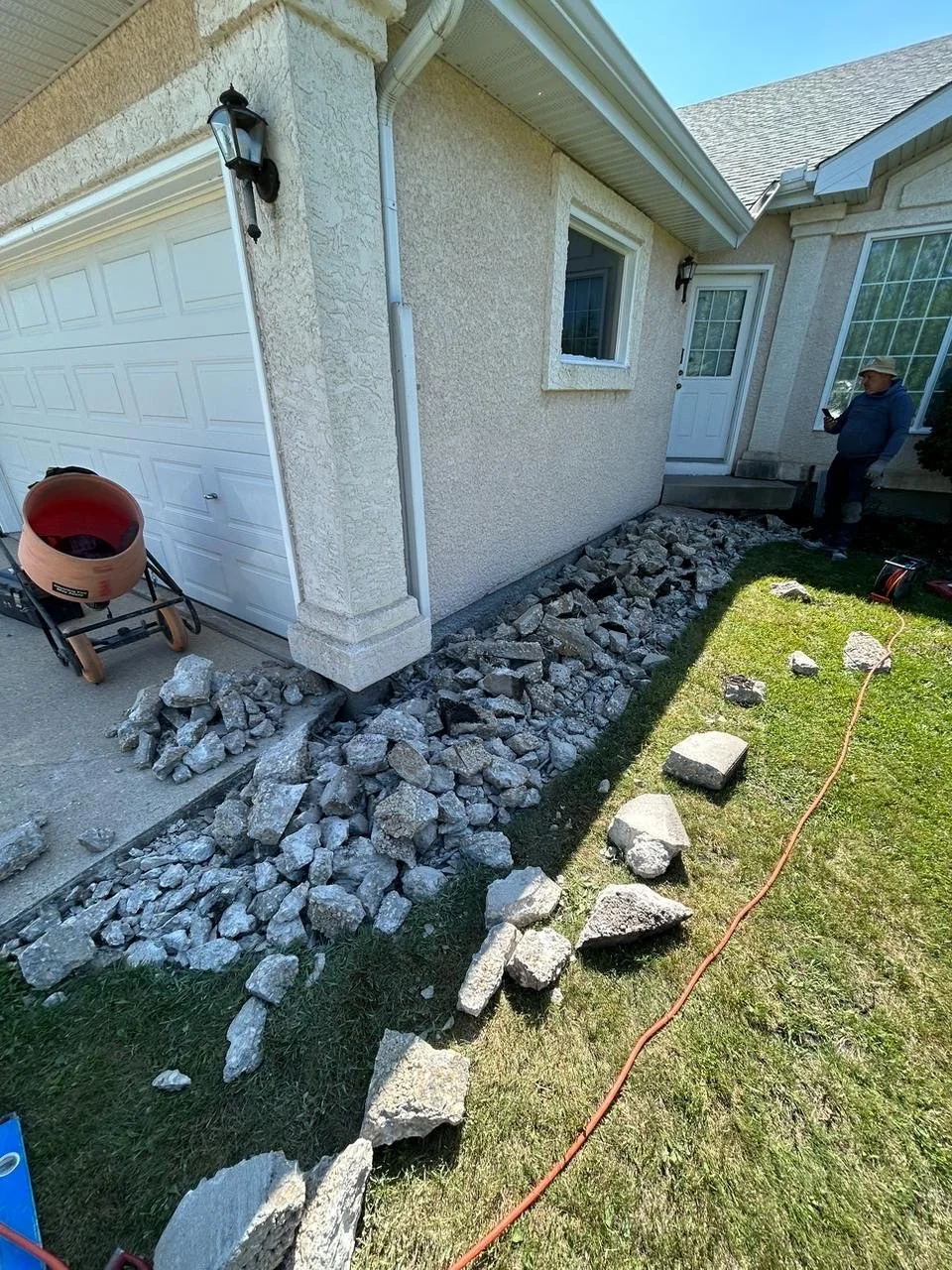 A person is working on landscaping or foundation work outside a house, with a pile of rocks and broken concrete along the side of the house near the grass. There is a white garage door and a white door with a window, as well as a window on the house.