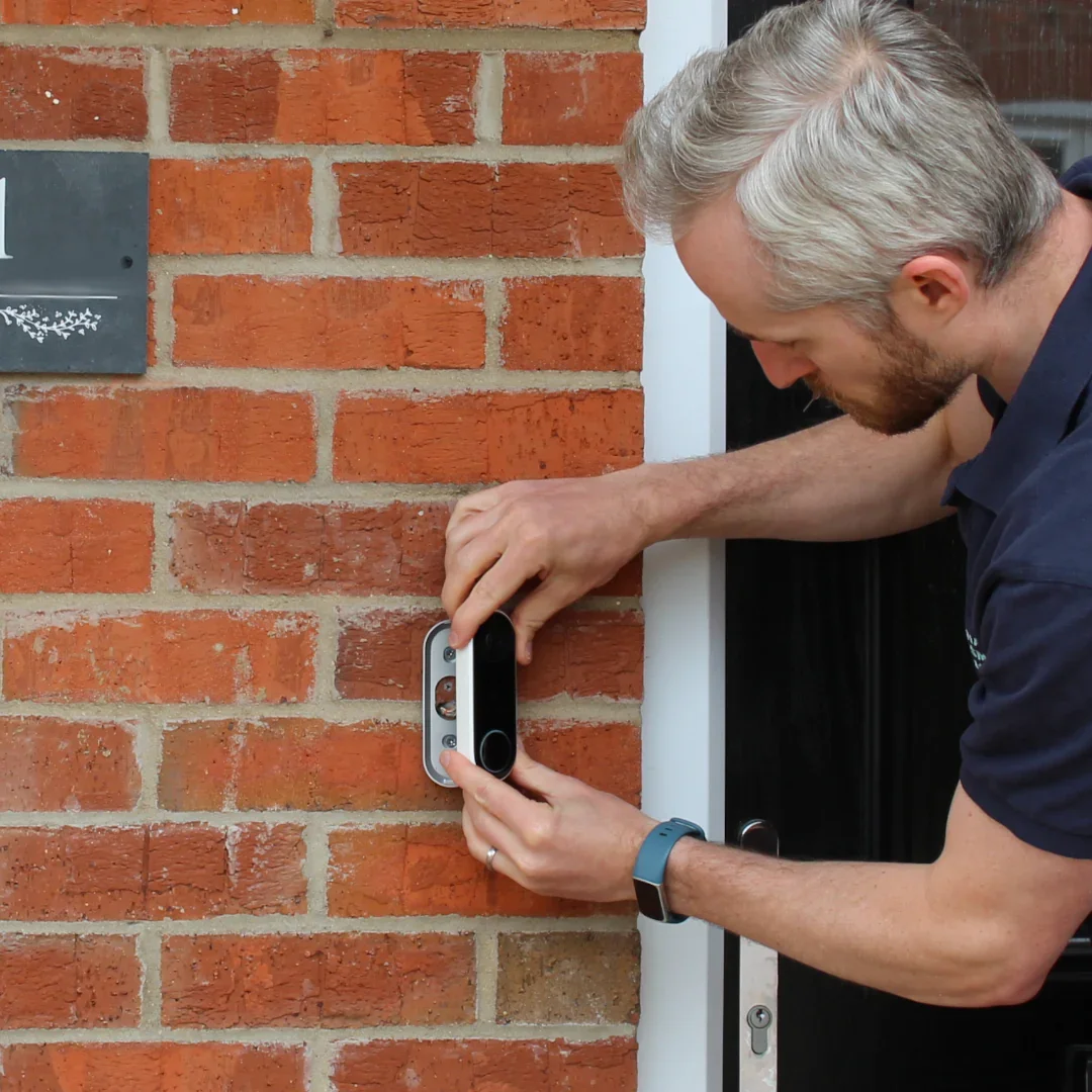 A man installing a smart doorbell or security camera on a brick wall outside a house.