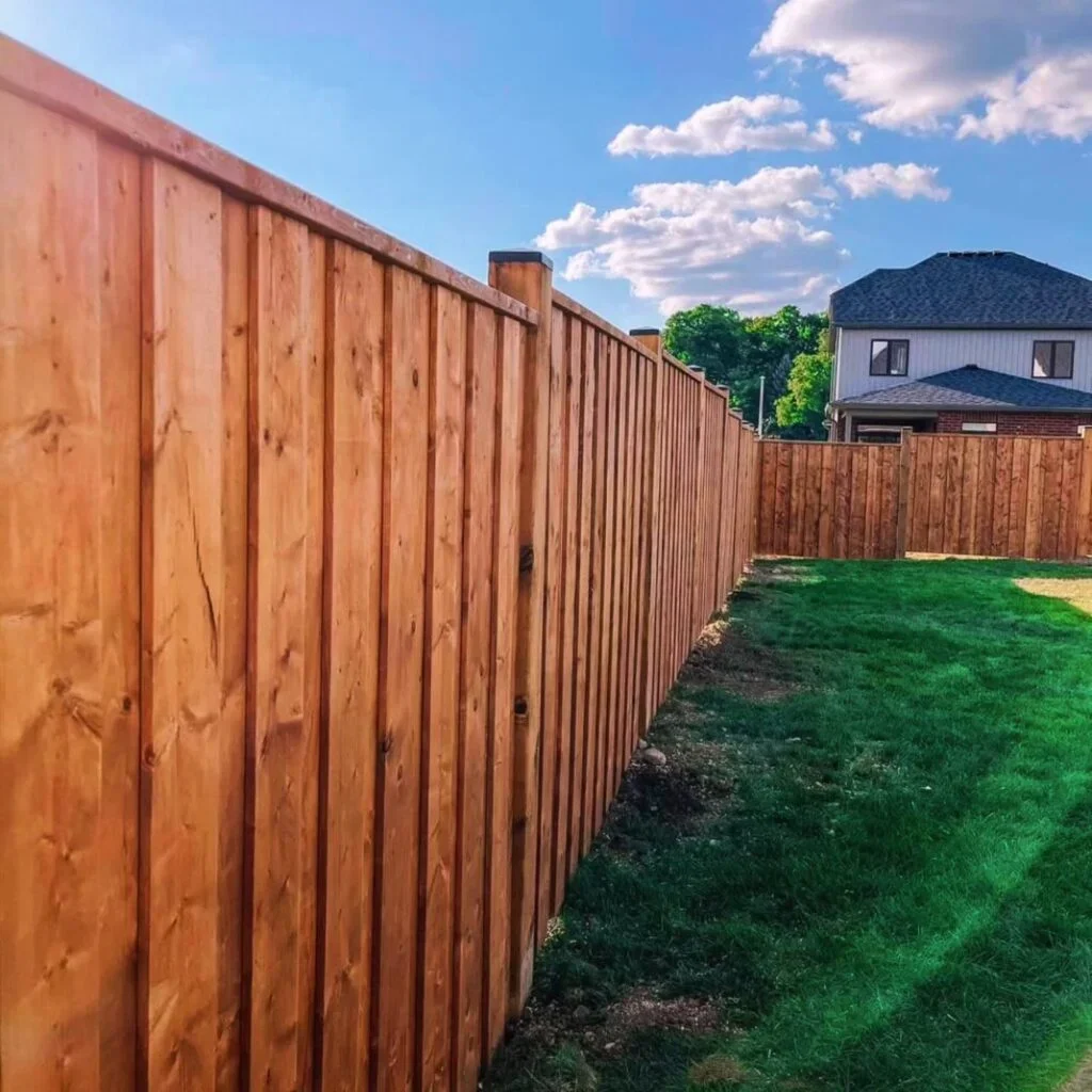 A newly installed wooden fence running along a green grassy backyard, with a house in the background and a partly cloudy sky.