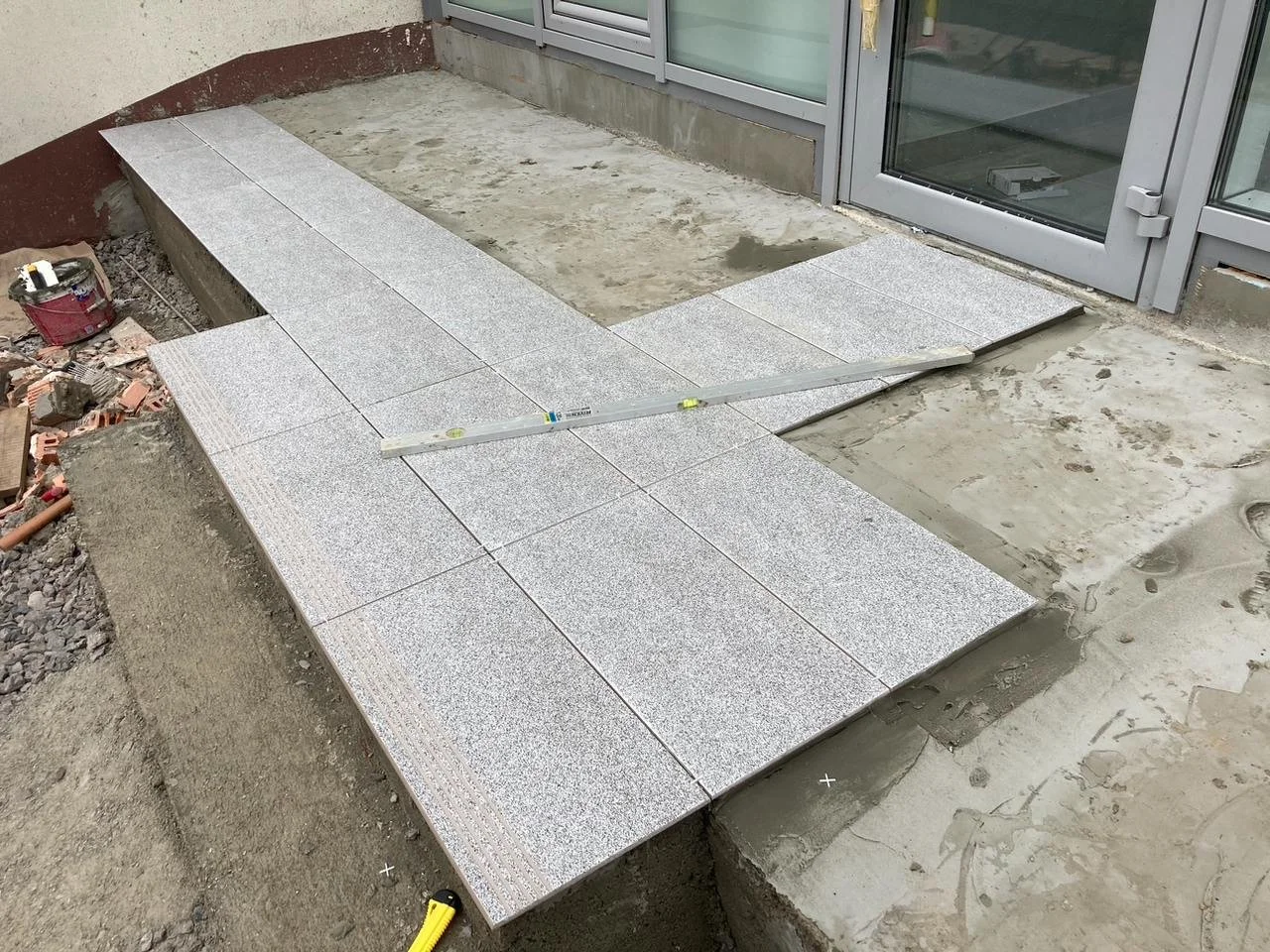 Outdoor construction site with grey tiled walkway being laid, glass door and sliding glass door in the background, and construction tools and materials scattered around.
