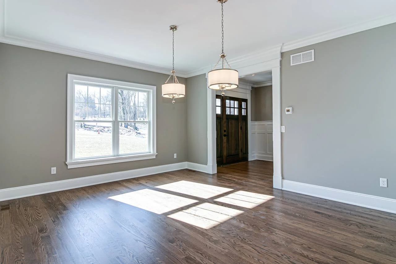 Empty living room with hardwood floors, large window, gray walls, white trim, and a front door.