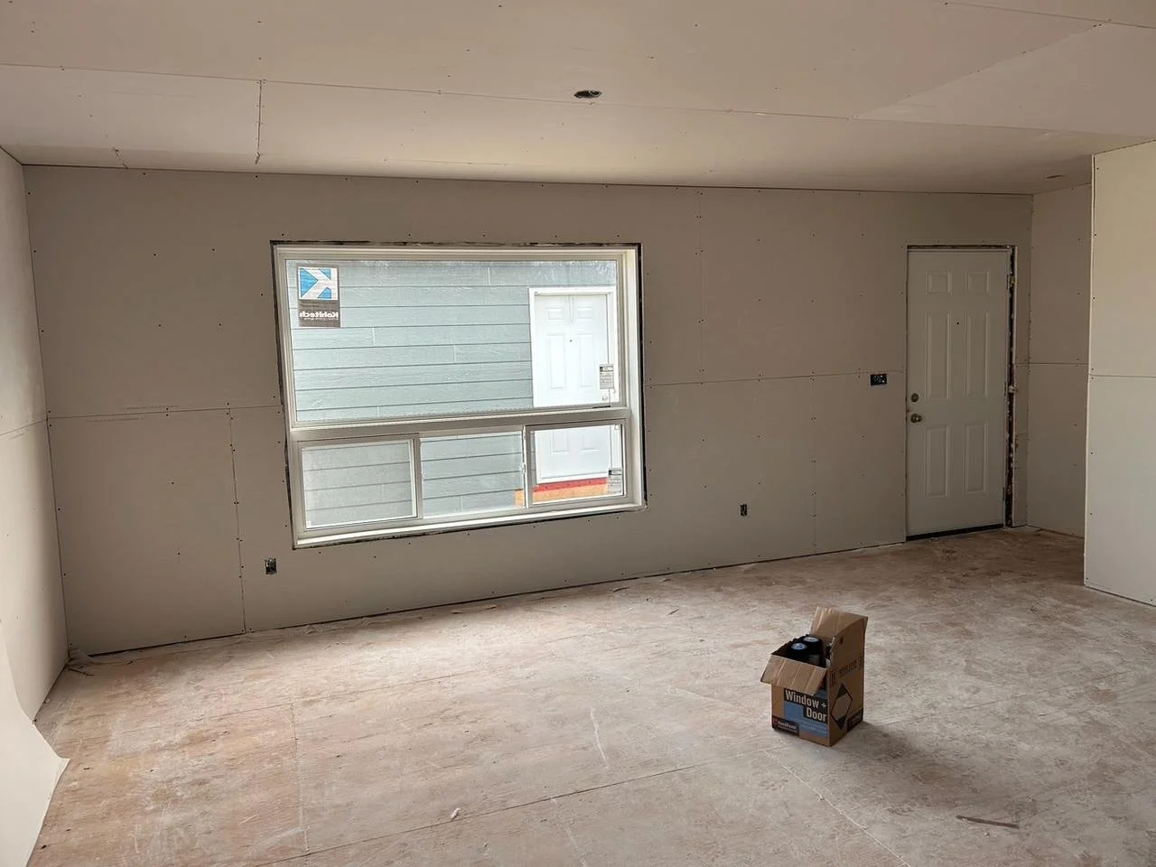 Empty room under construction with unfinished drywall, a window showing the outside, and a door. There is a cardboard box on the floor containing paint supplies.