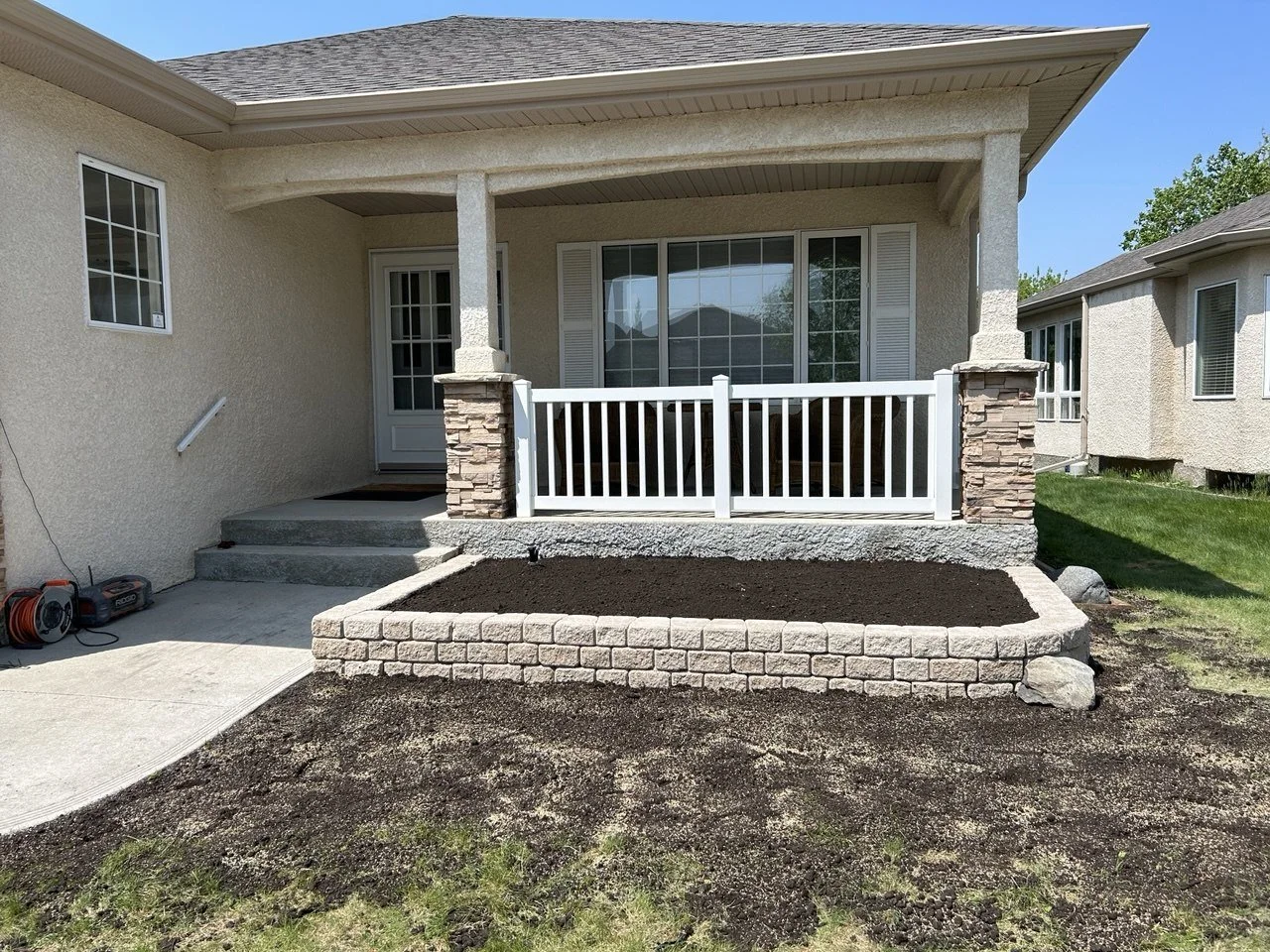 Backyard porch with a white railing, stone pillars, and a small garden bed in front, with freshly tilled soil, neighboring houses, green grass, and clear blue sky.