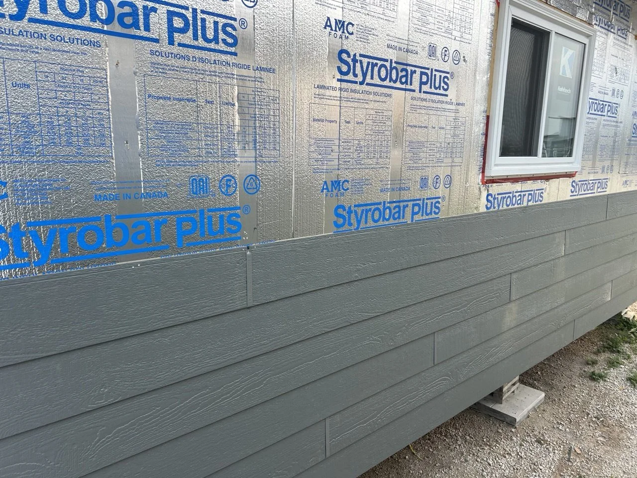 Exterior wall of a building under construction, with gray siding and insulated sheathing labeled 'Styrobar Plus' and 'AMC Foam', and a white-framed window installed.