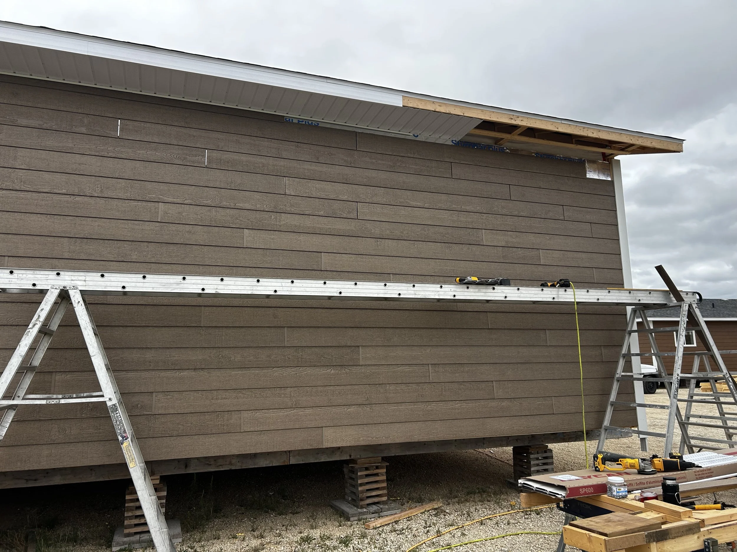 Construction site where a house's exterior wall is being installed with brown siding. An unfinished roof overhang is visible, with construction tools on a workbench and scaffolding nearby, under a cloudy sky.