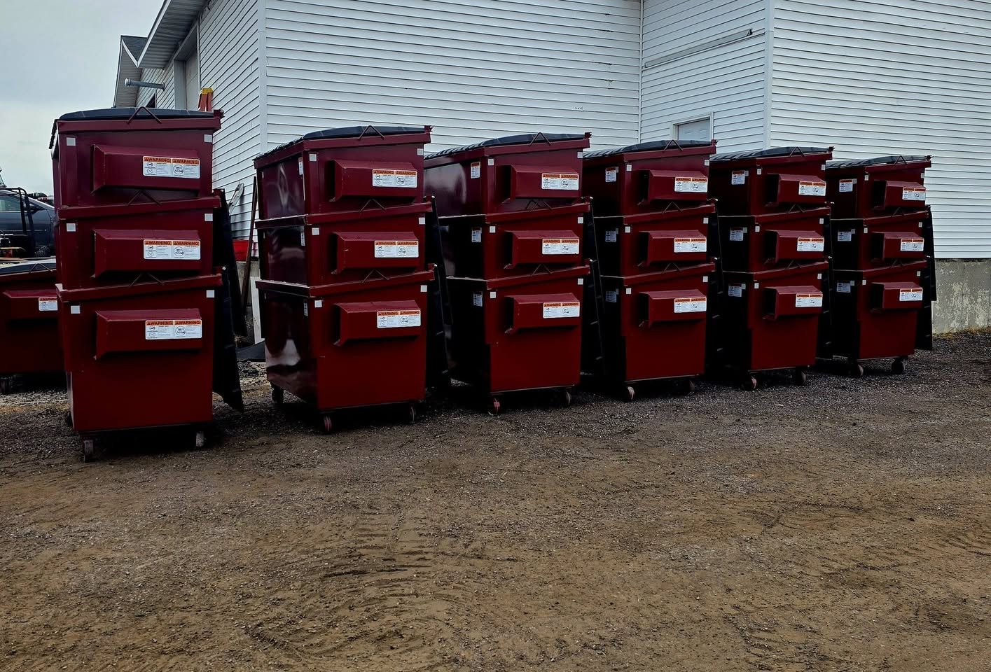 Multiple red dumpsters lined up outside near a white building with bushed in the background.
