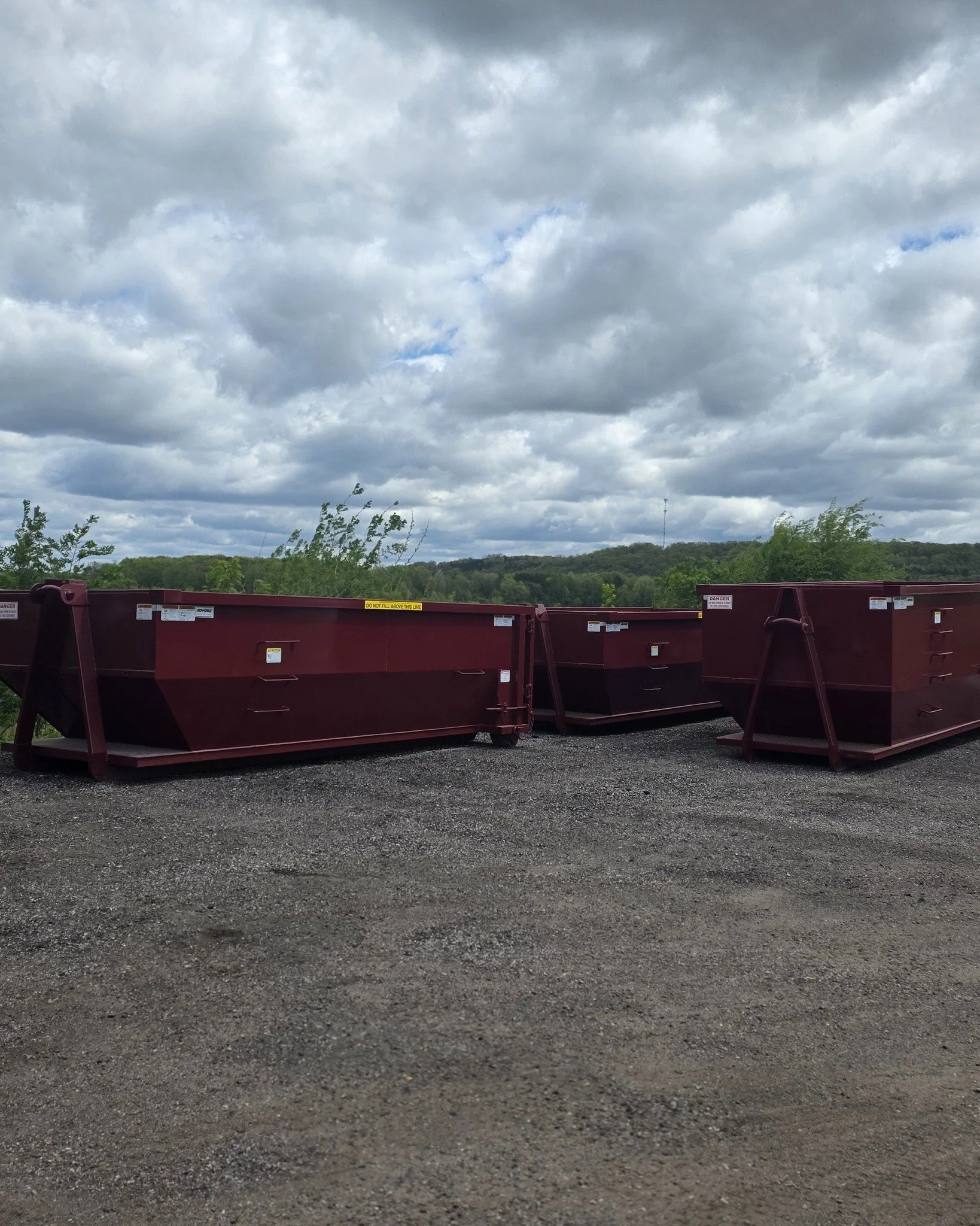 Overcast sky with gray clouds over a landscape of green trees and rolling hills, with a foreground of dark gravel and red metal equipment or barriers.