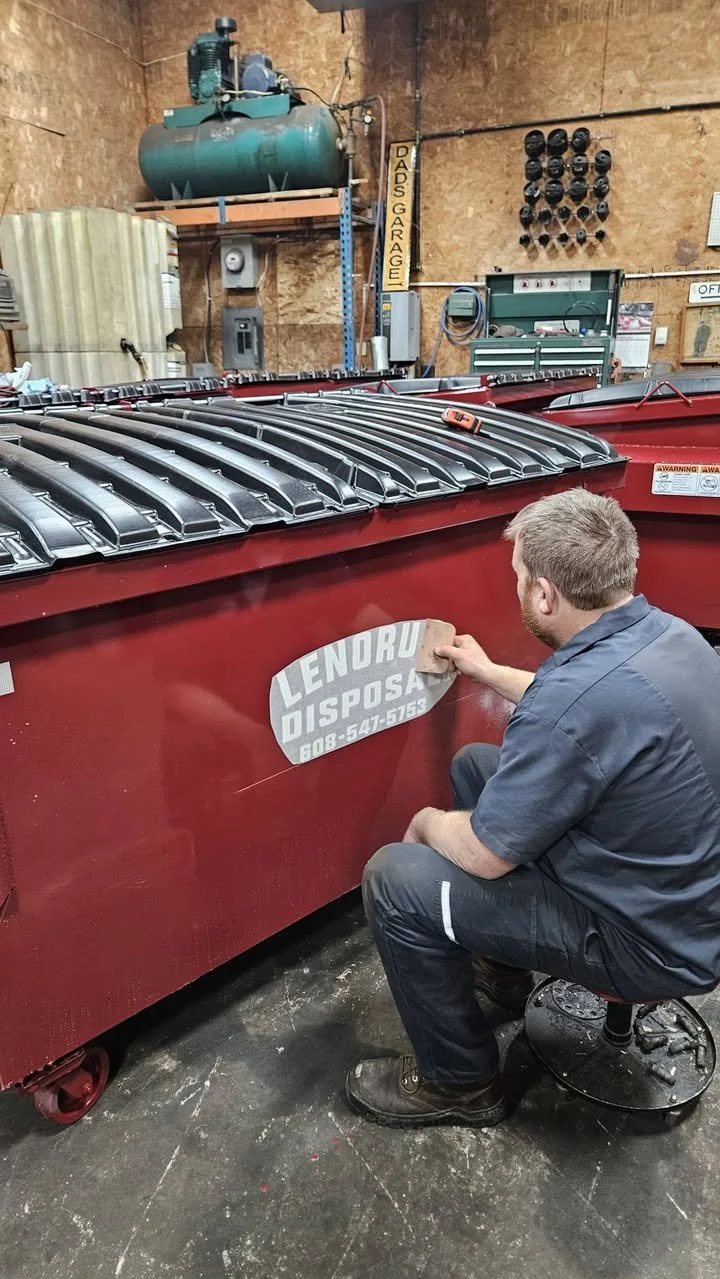 A man kneeling on a mechanic stool in a garage, applying a decal that reads 'Lendru Disposal' with a phone number onto a large red industrial dumpster. The garage has wooden walls, various tools, and a sign that says 'Dads Garage'.