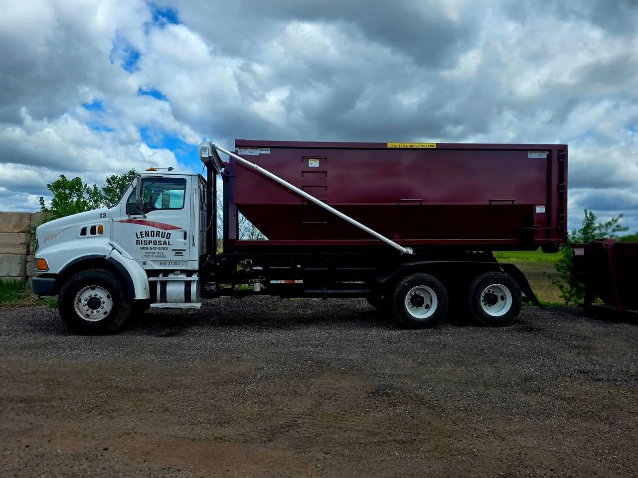 A white dump truck with maroon container parked on dirt ground under cloudy sky.