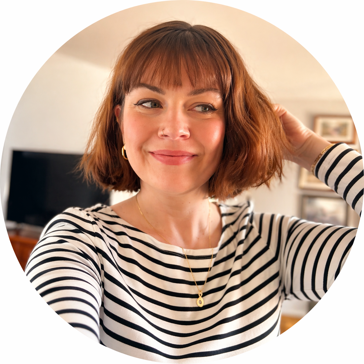 A woman with short, wavy, auburn hair smiling and adjusting her hair with one hand. She is wearing a black and white striped shirt, gold hoop earrings, a gold necklace, and a nose ring. The background is a cozy living room with a television and framed pictures on the wall.