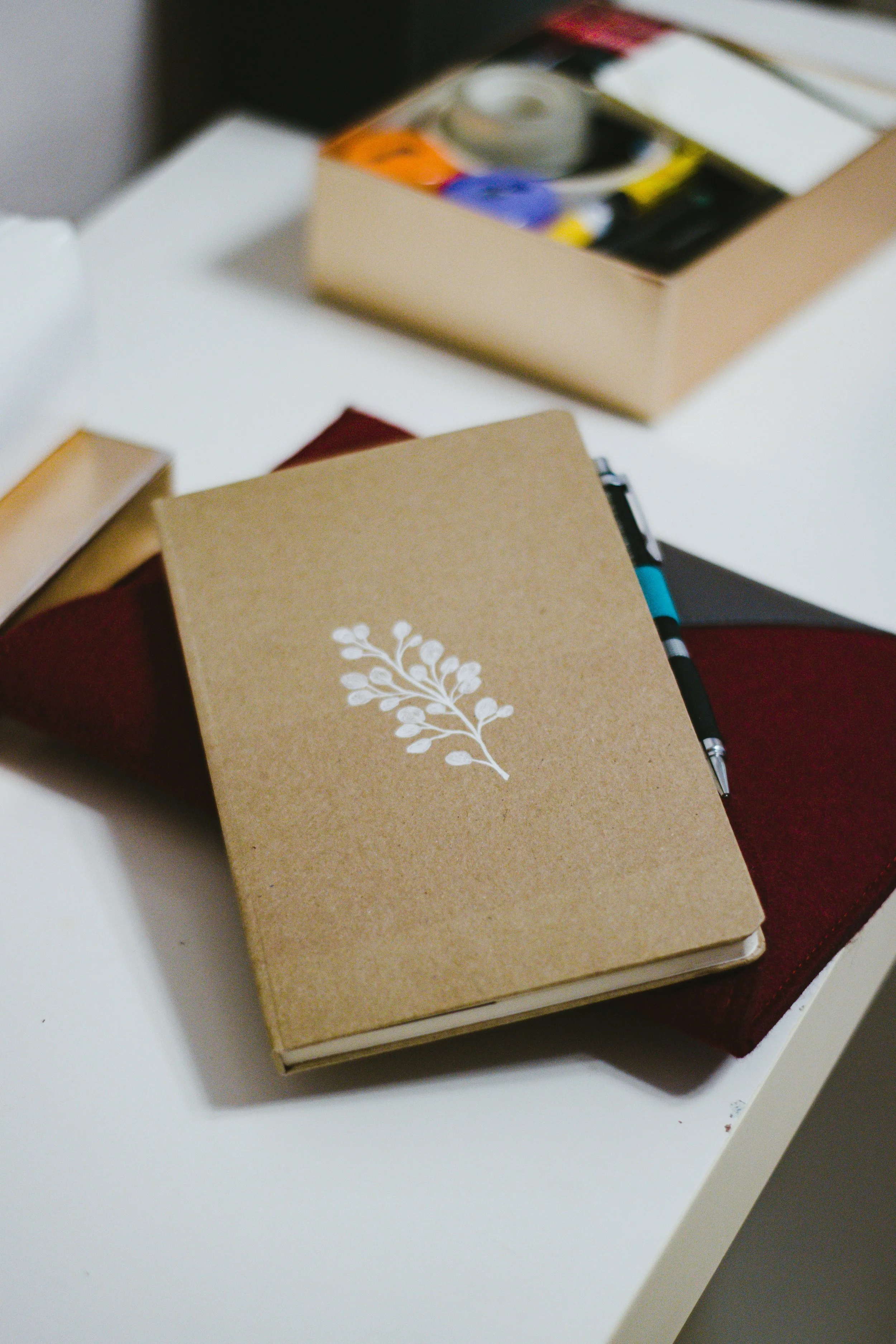 Various notebooks and a pen on a white table, with a book featuring a white floral design on a tan cover.