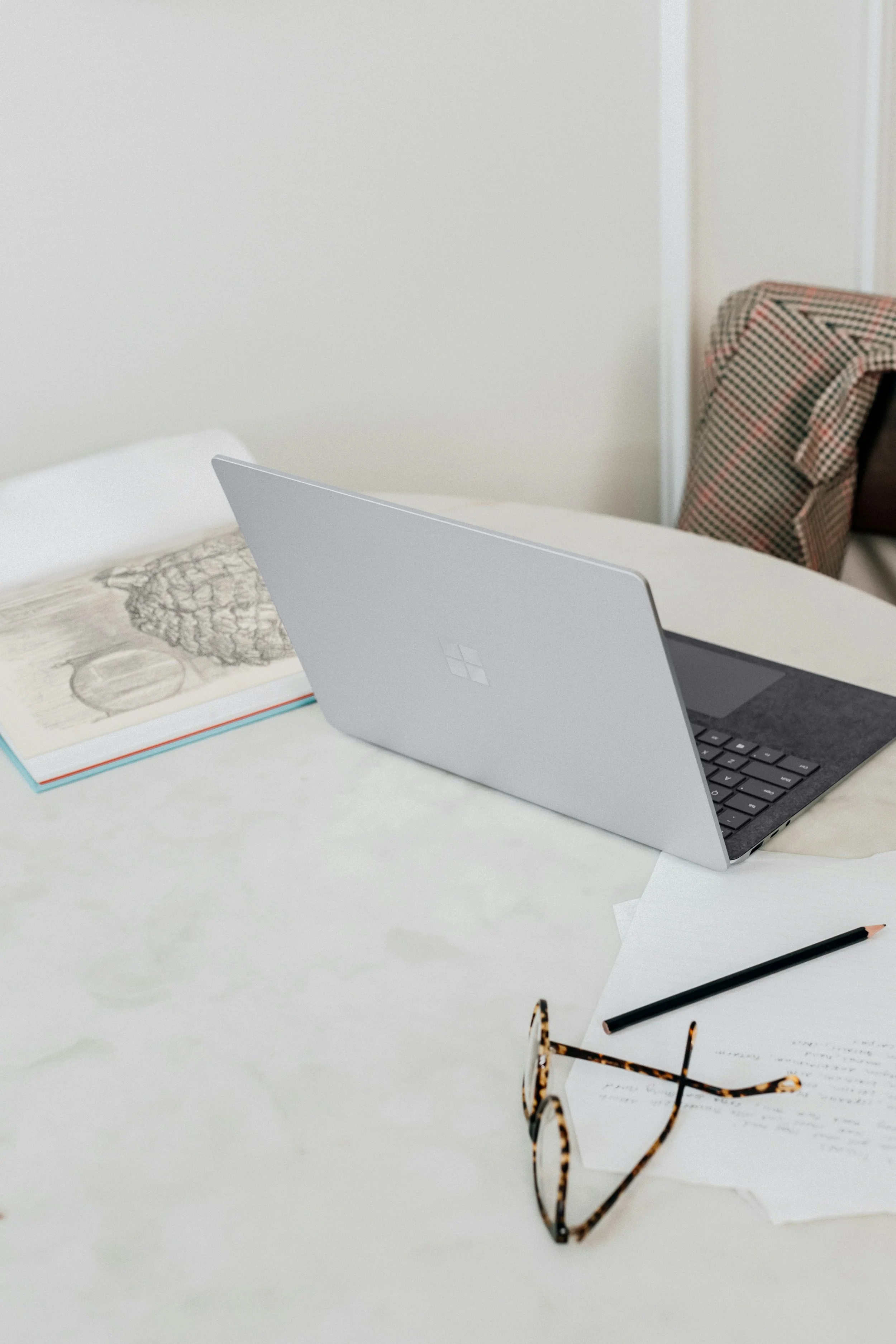 A laptop on a white desk with scattered papers, a pair of tortoiseshell glasses, a black pencil, and a pair of glasses with tortoiseshell frame, in a workspace with a patterned chair in the background.