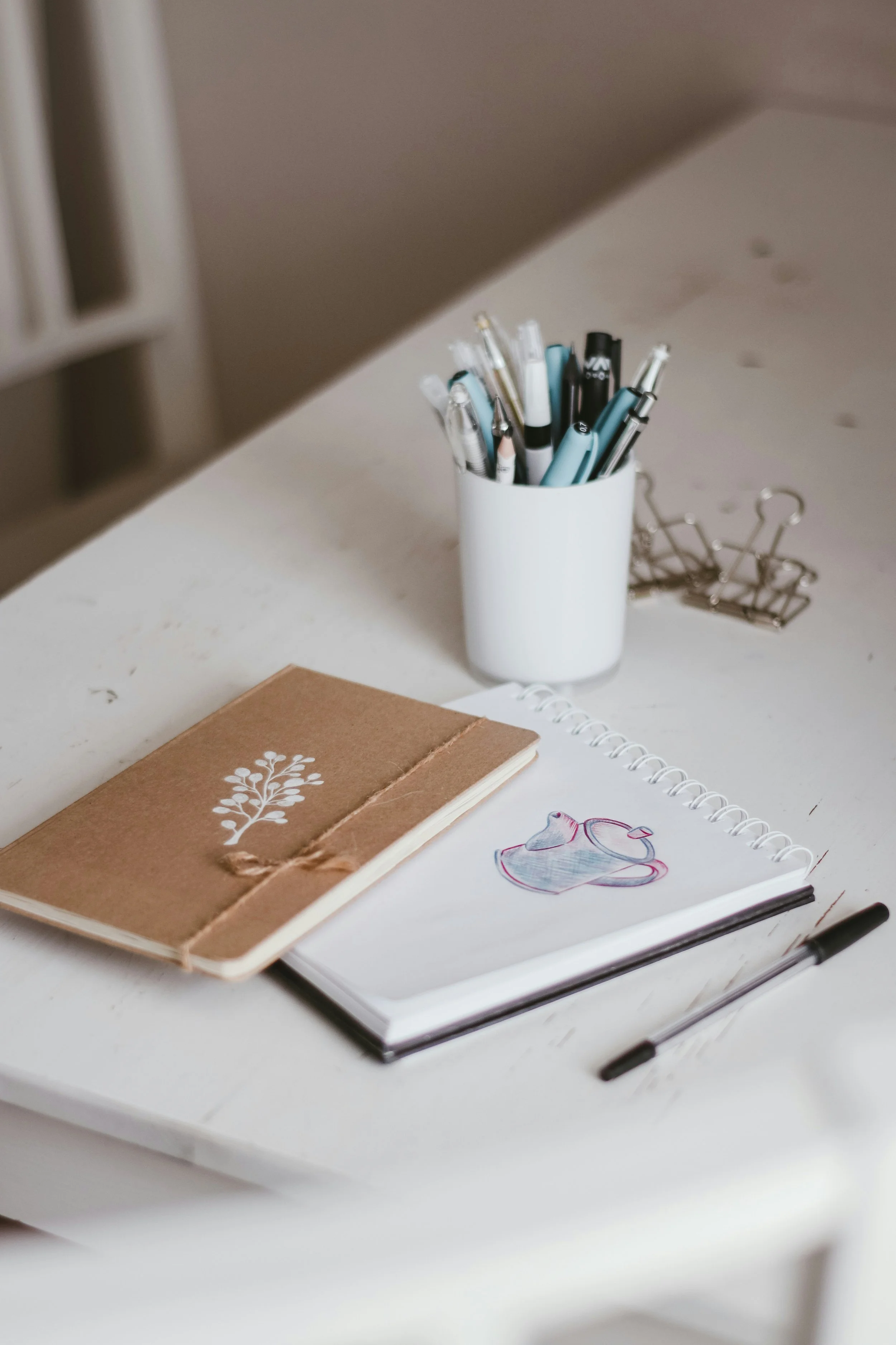 A white table with a brown notebook, a notebook with a drawing of a teapot, a spiral notebook, black pens, a cup holding pens and markers, and a black paperclip.