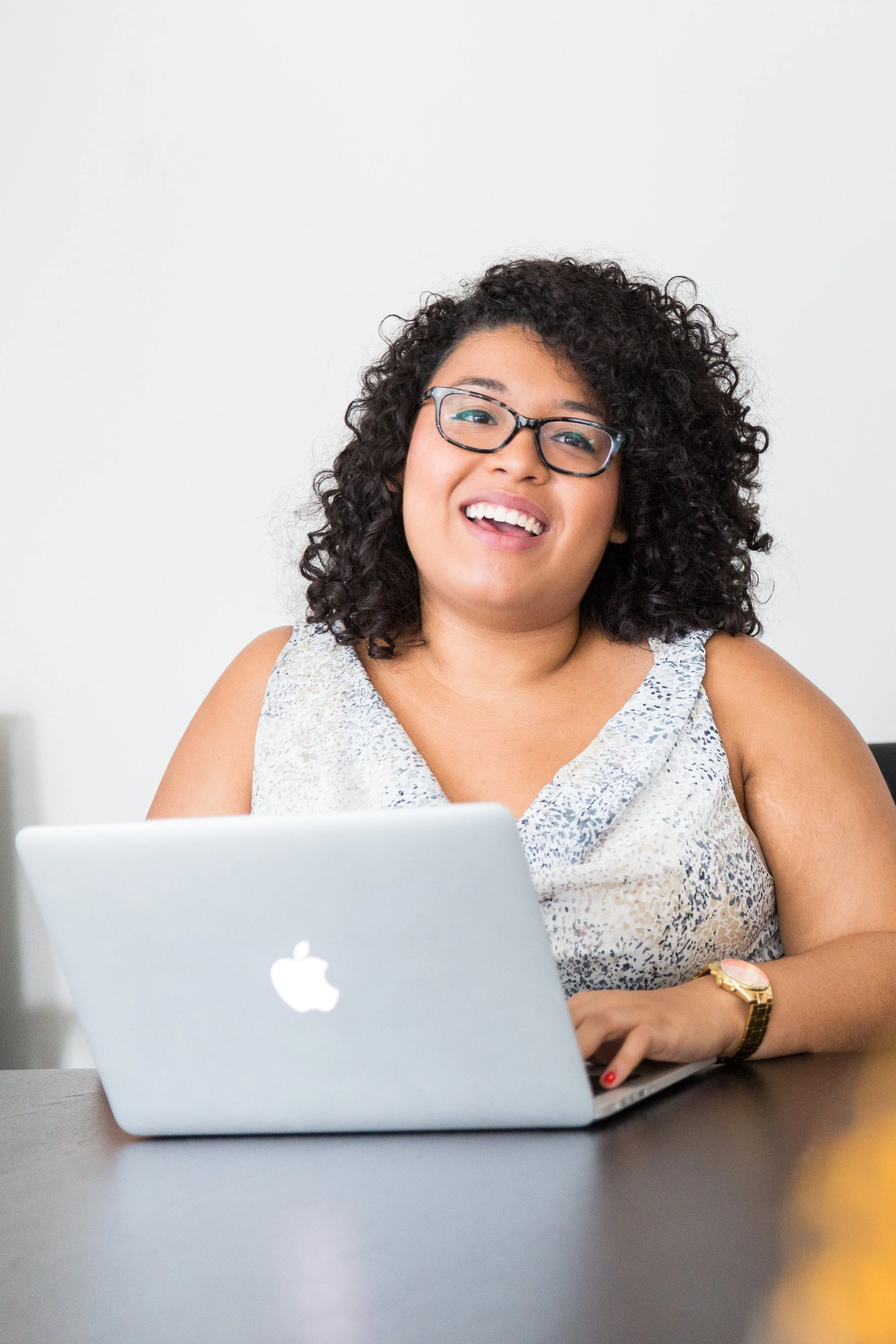 A woman with curly black hair, glasses, and a sleeveless patterned top sitting at a table with a silver MacBook laptop, smiling at the camera against a plain white background.
