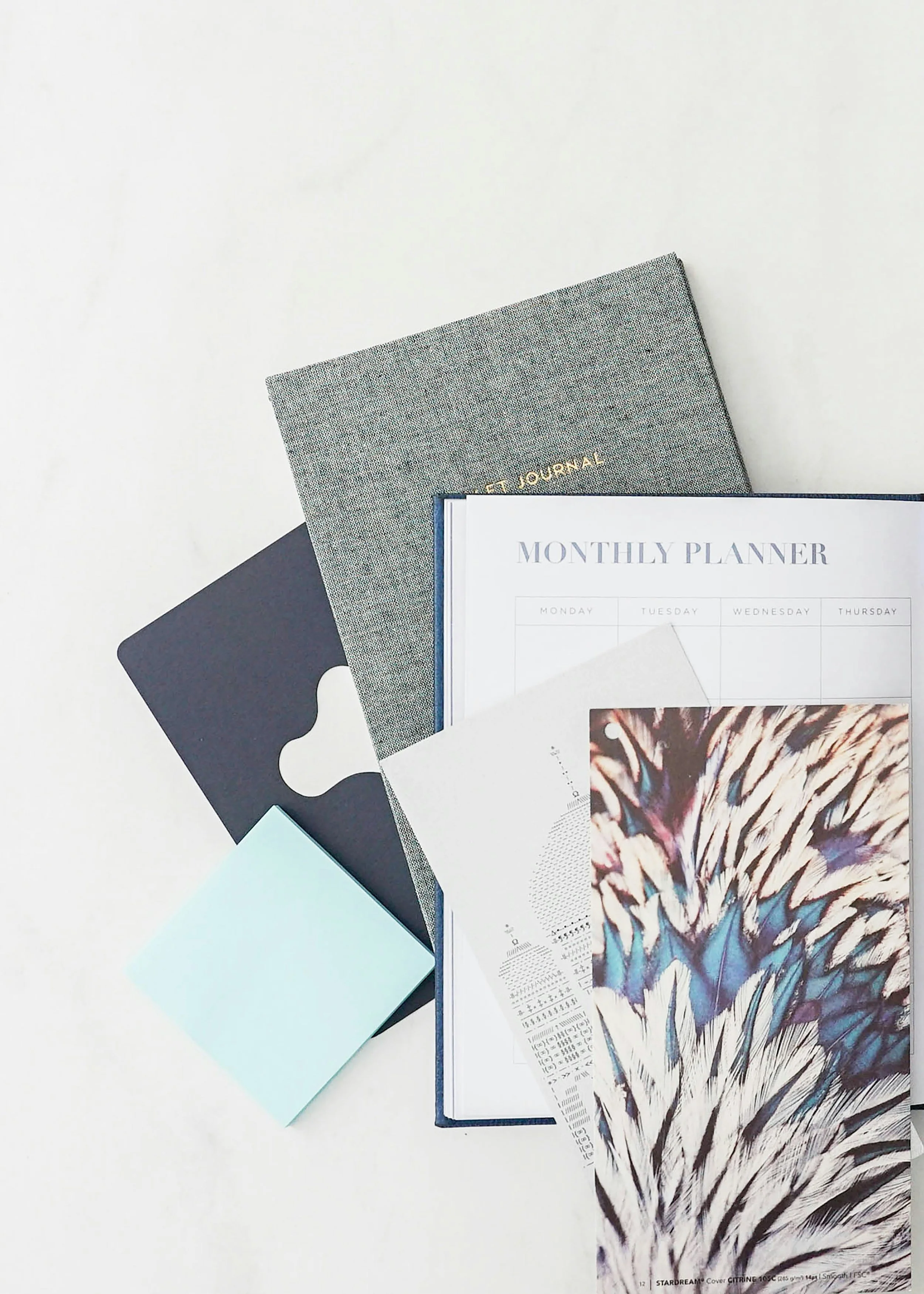 A flat lay of various stationery items including a gray textured notebook with gold lettering, a black paper clip, a monthly planner with a weekly layout, a receipt, a colorful printed paper, and a light blue sheet of paper, all arranged on a white surface.