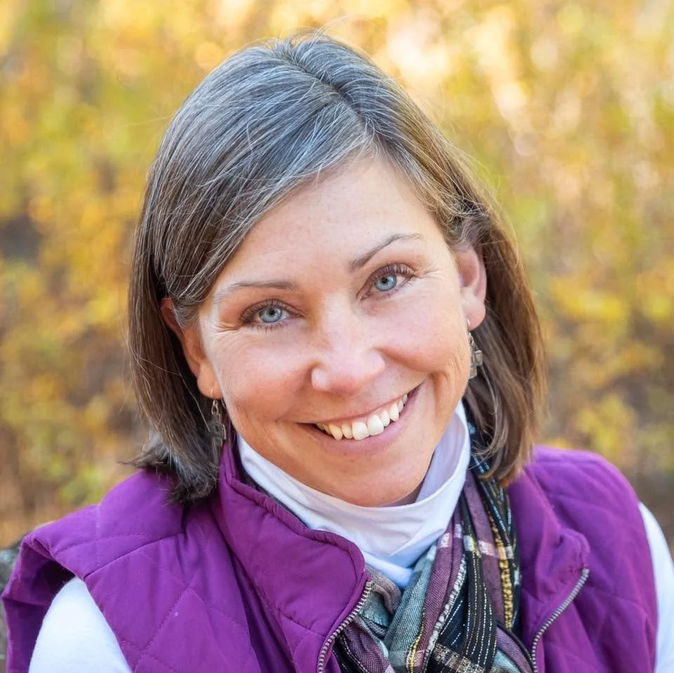 A woman with shoulder-length gray hair, blue eyes, and fair skin smiling outdoors with a background of autumn foliage. She is wearing a purple vest, a white shirt, and a scarf.