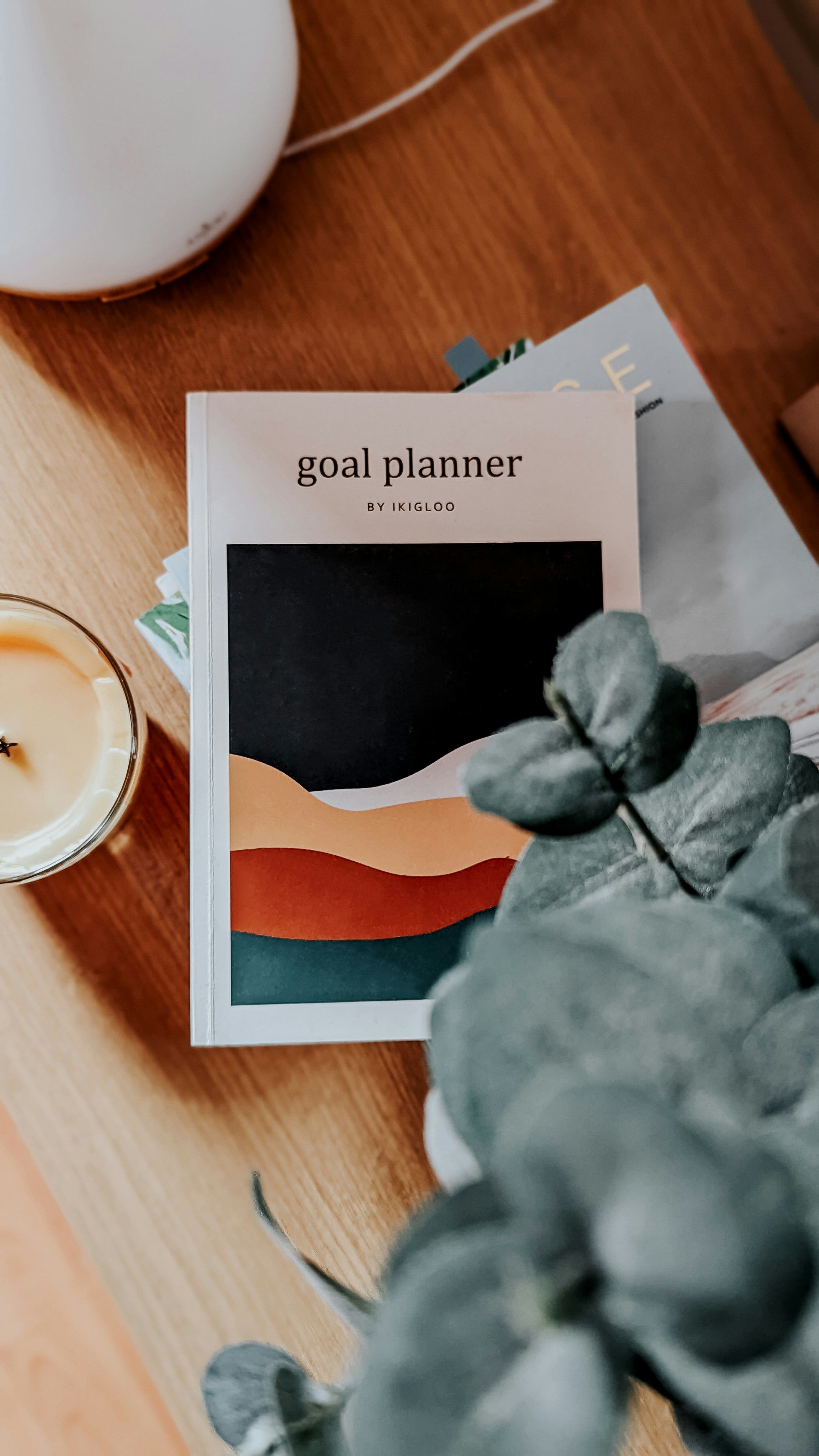 A goal planner book titled 'goal planner by ikigloo' with a colorful abstract design on the cover, a candle, and a plant on a wooden surface.