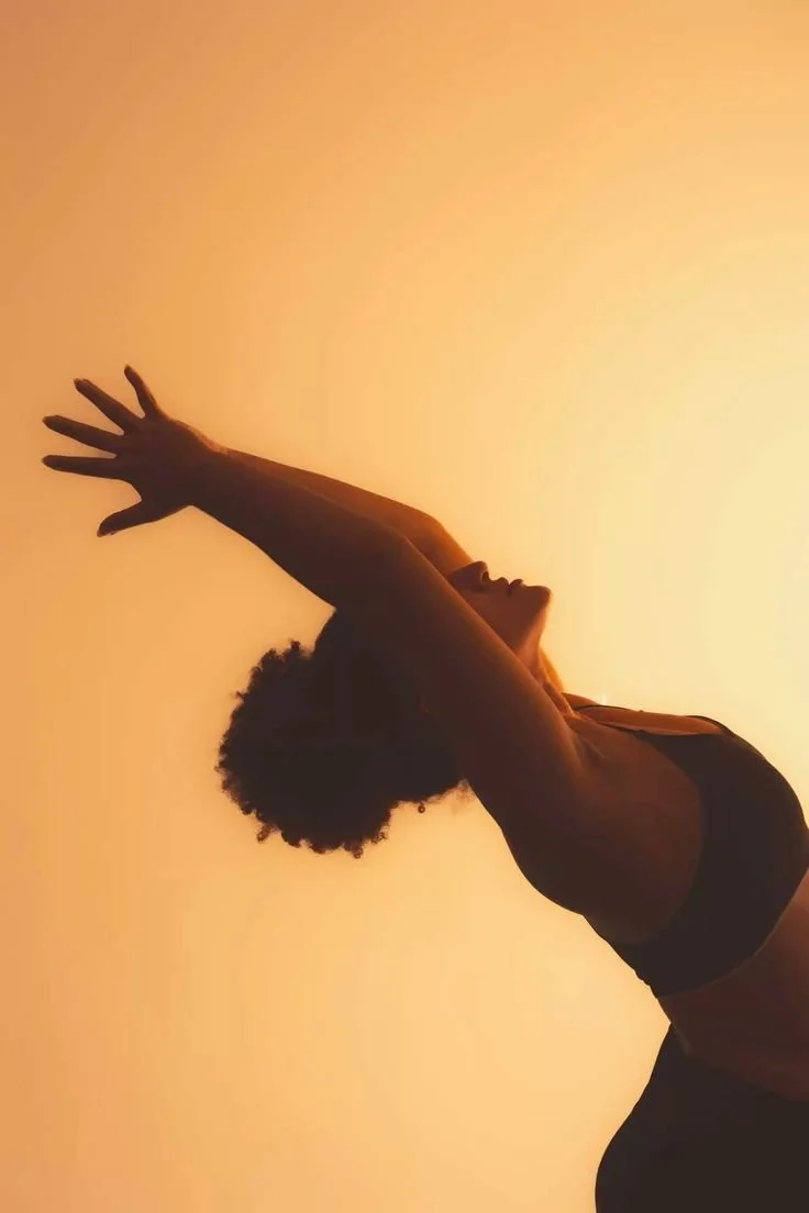 Persona realizando una pose de yoga o meditación con los brazos extendidos hacia arriba, contra un cielo al atardecer o amanecer.