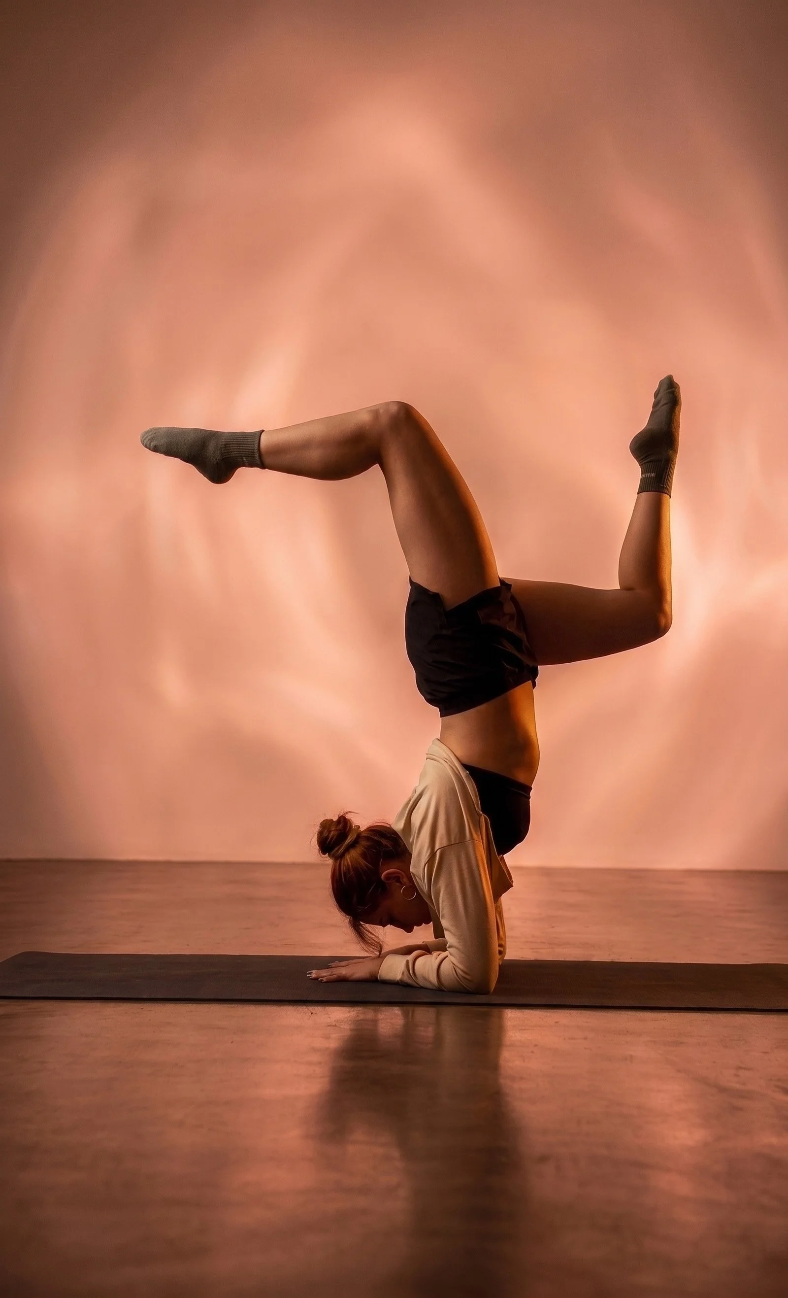 Una mujer haciendo una postura de yoga en el suelo, con las piernas elevadas y formando un ángulo recto, en un estudio con paredes rosadas.