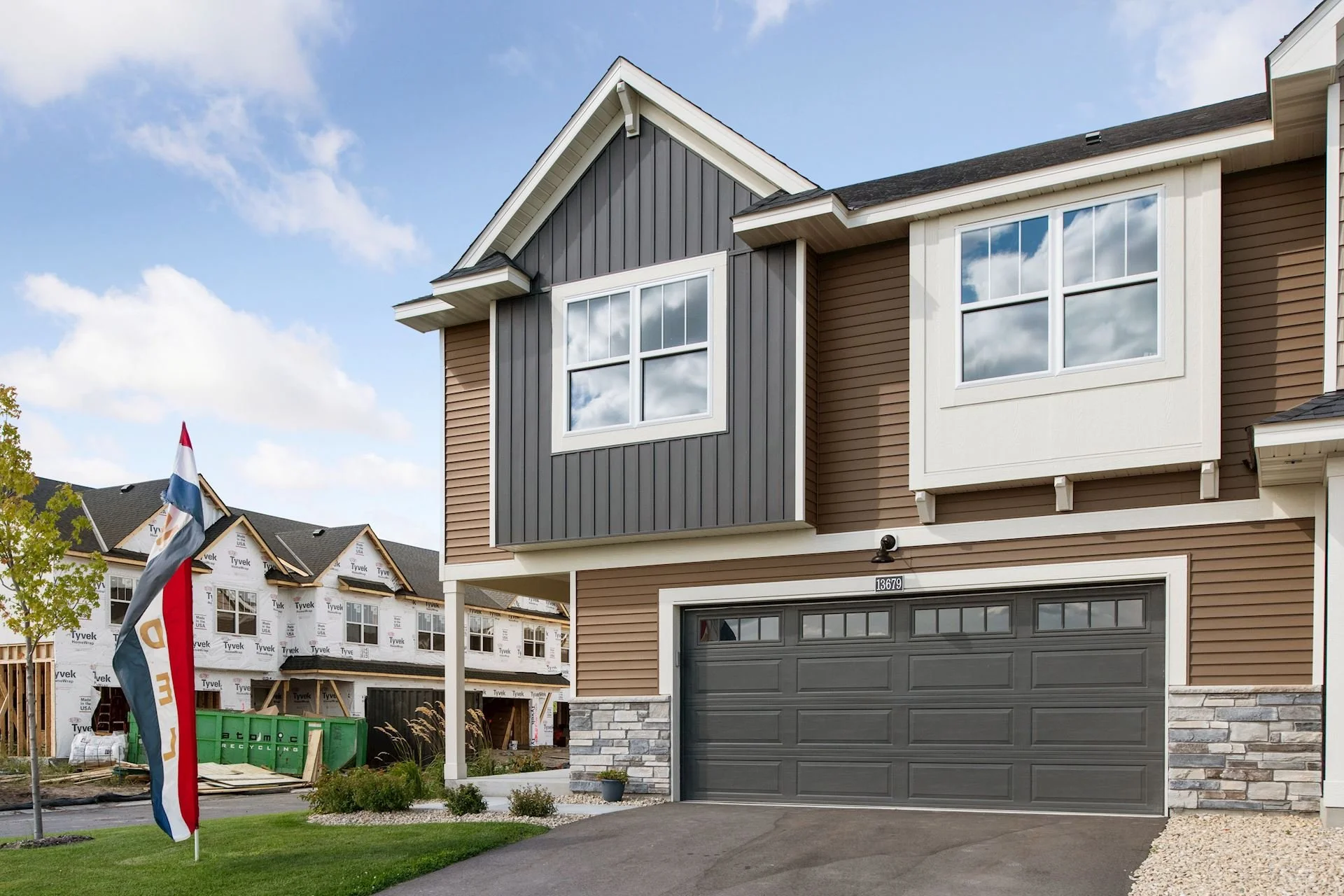 Newly constructed two-story house with tan and dark gray exterior, double garage, and large windows. Construction ongoing on a neighboring house in background with visible building materials. Flag of the Netherlands in front yard.