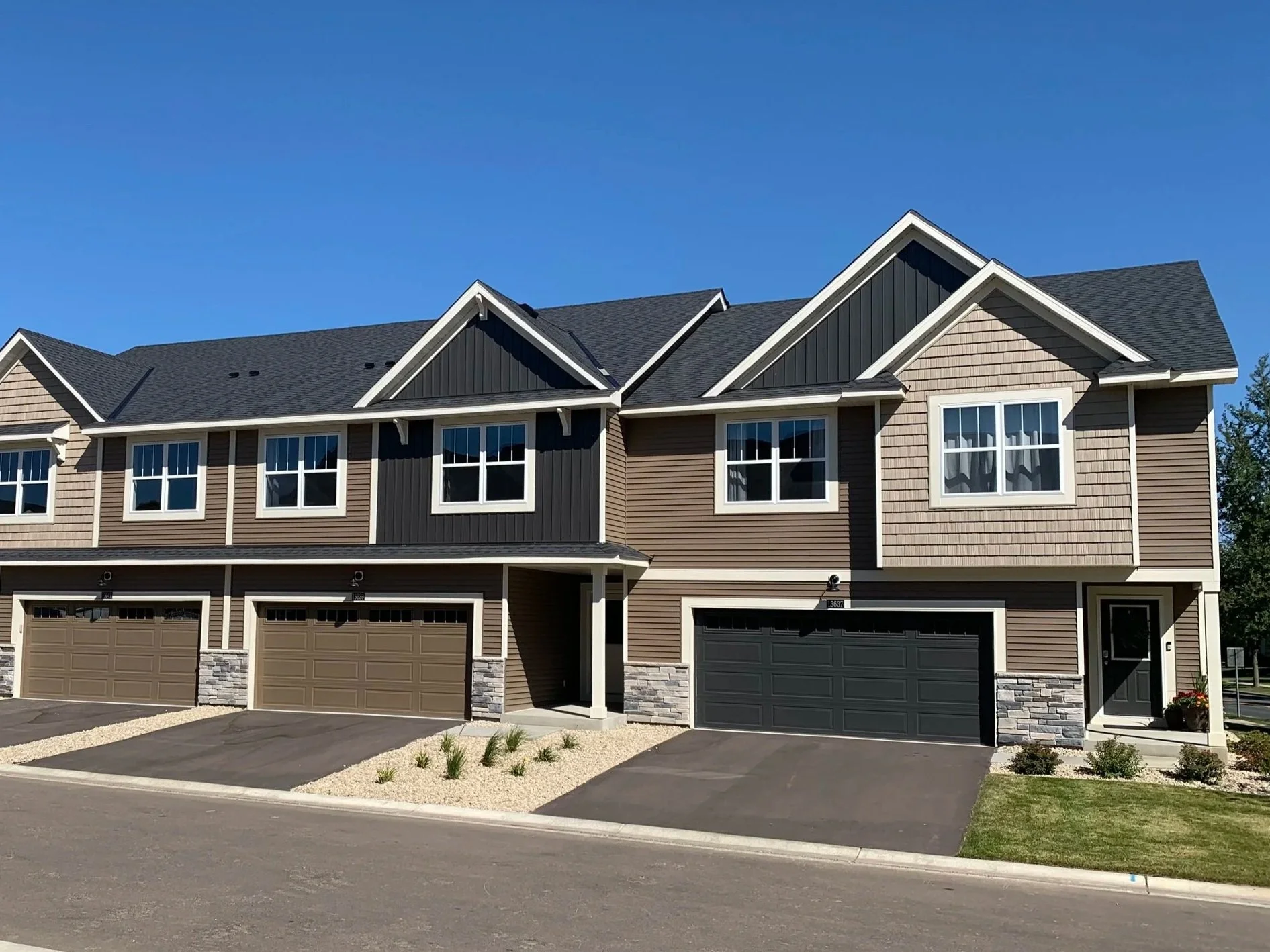 Multiple attached townhouses with garage doors on the ground level, beige and dark gray siding, peaked roofs, and large windows. Clear blue sky above and small landscaped areas in front.