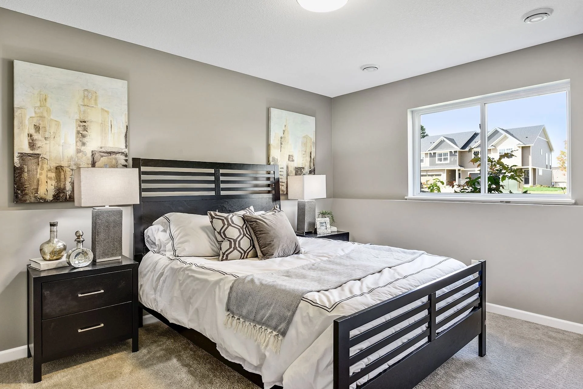 A bedroom with a black metal bed frame, white bedding, and decorative pillows. Two nightstands with lamps and decorative items. Two abstract paintings above the bed. A large window showing neighboring houses and greenery outside.