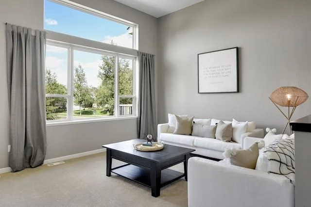Living room with large window, white sofa, black coffee table, beige pillows, black side table with lamp, and framed artwork on gray wall.