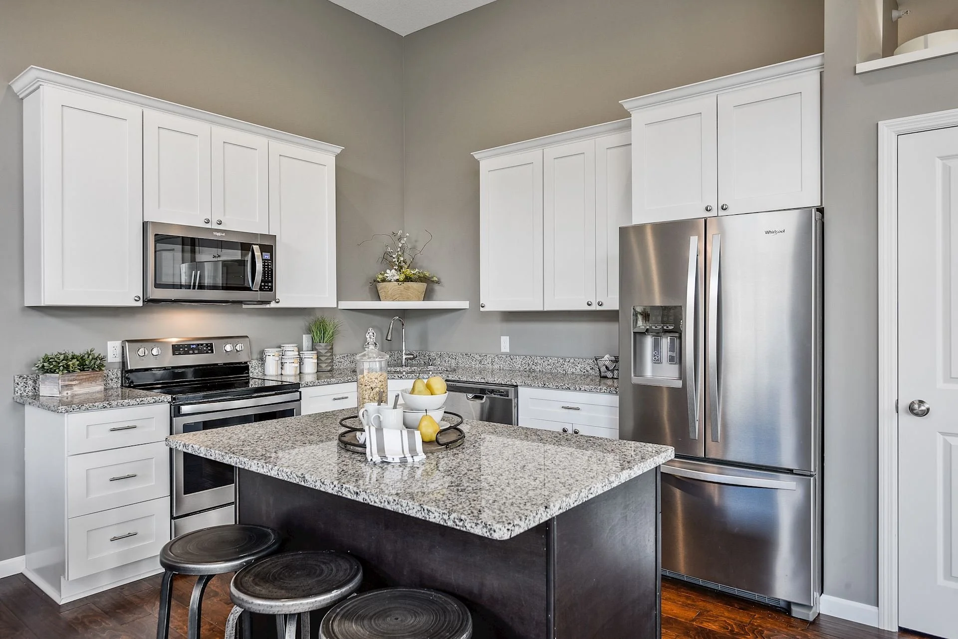 A modern kitchen with white cabinets, stainless steel appliances, and a granite island. The countertop has a bowl of pears, a jar of popcorn, and a tray with cups and a napkin. There are three black stools at the island and a small potted plant on th