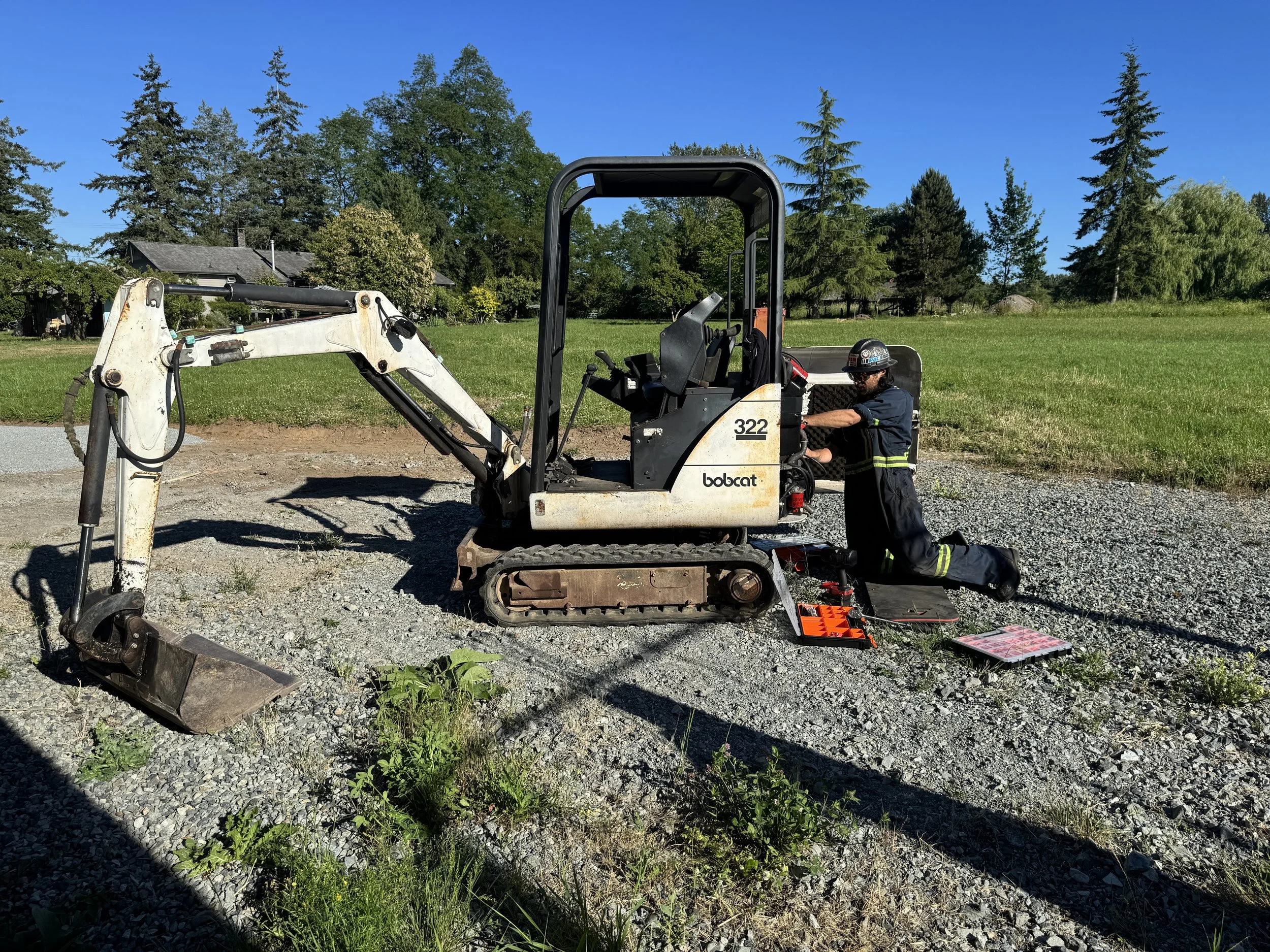 A construction worker kneeling beside a Bobcat mini excavator and inspecting it, with tools and equipment laid out nearby, on a gravel surface with grassy area and trees in the background.