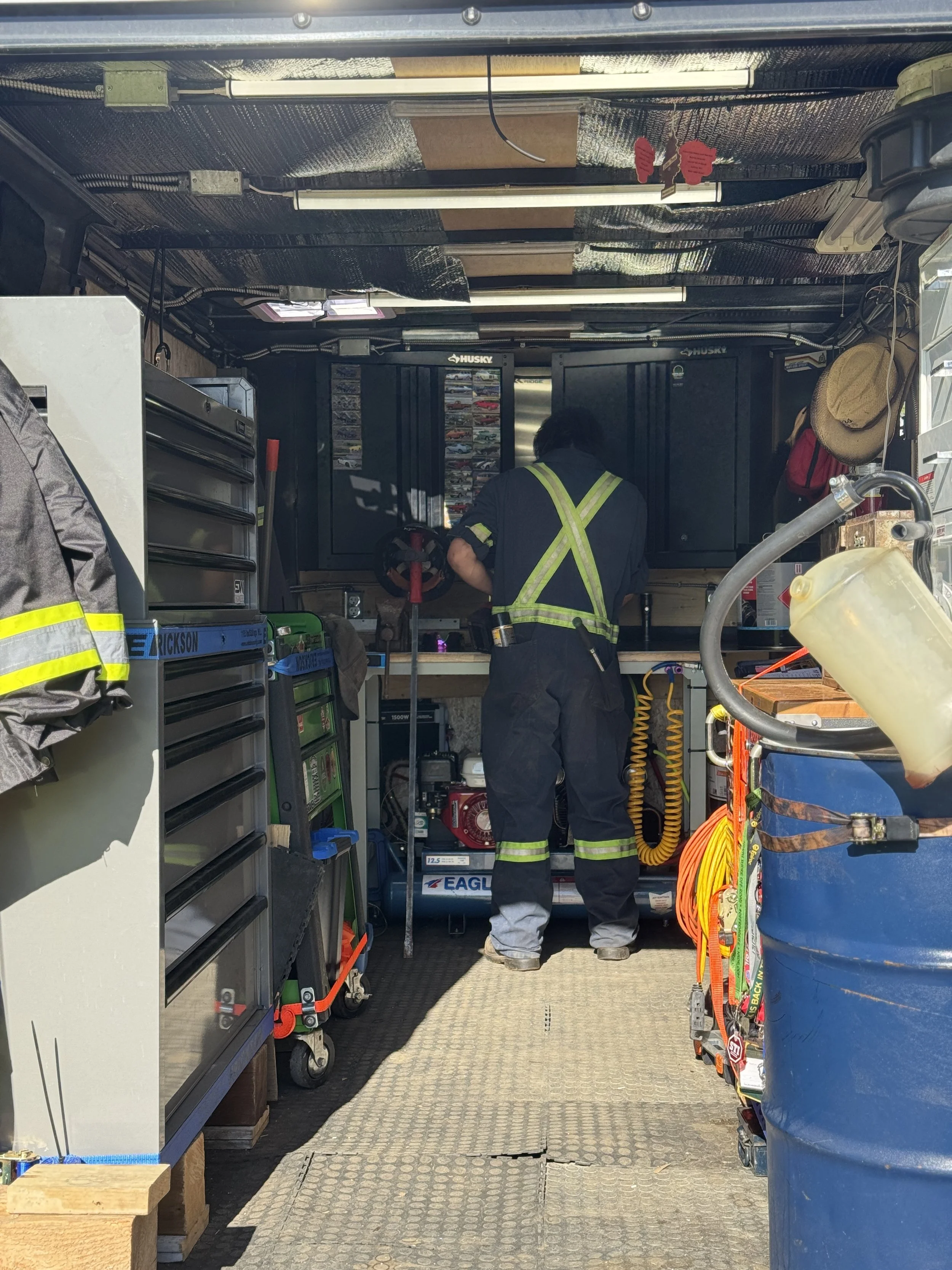 A mechanic in coveralls inside a utility trailer, surrounded by tools and supplies, with various equipment on the ceiling and shelves, and a large blue barrel on the right side.