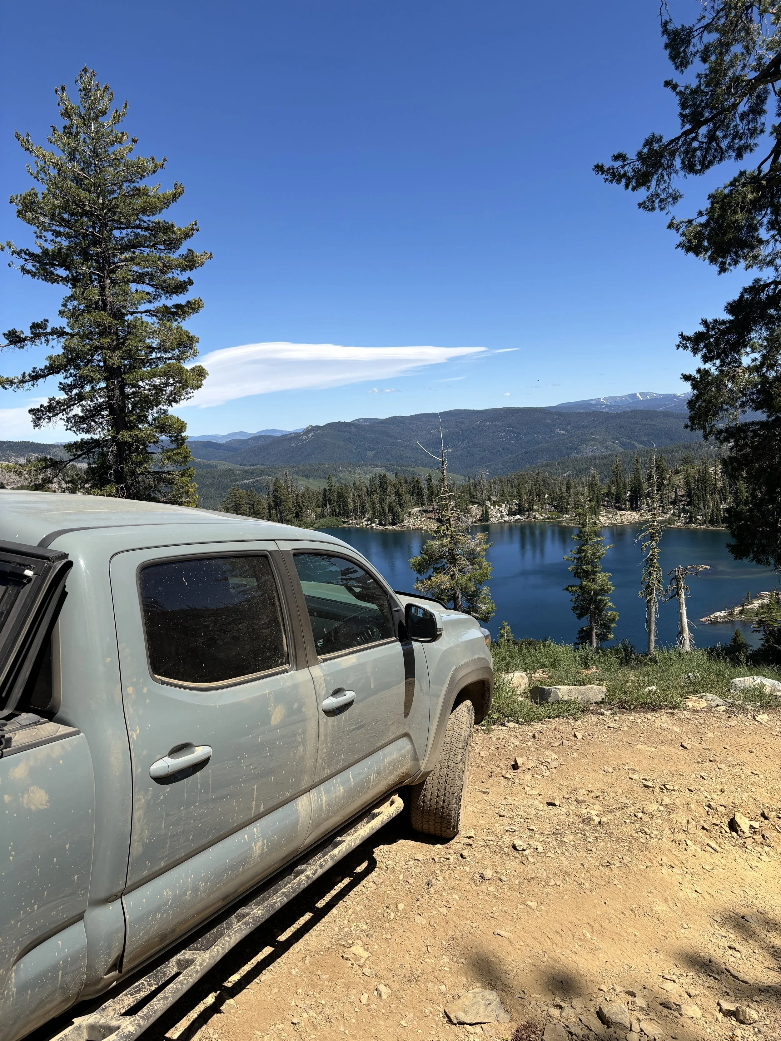 A silver pickup truck parked on a dirt road in front of a lake surrounded by pine trees and mountain ranges under a clear blue sky with some clouds.