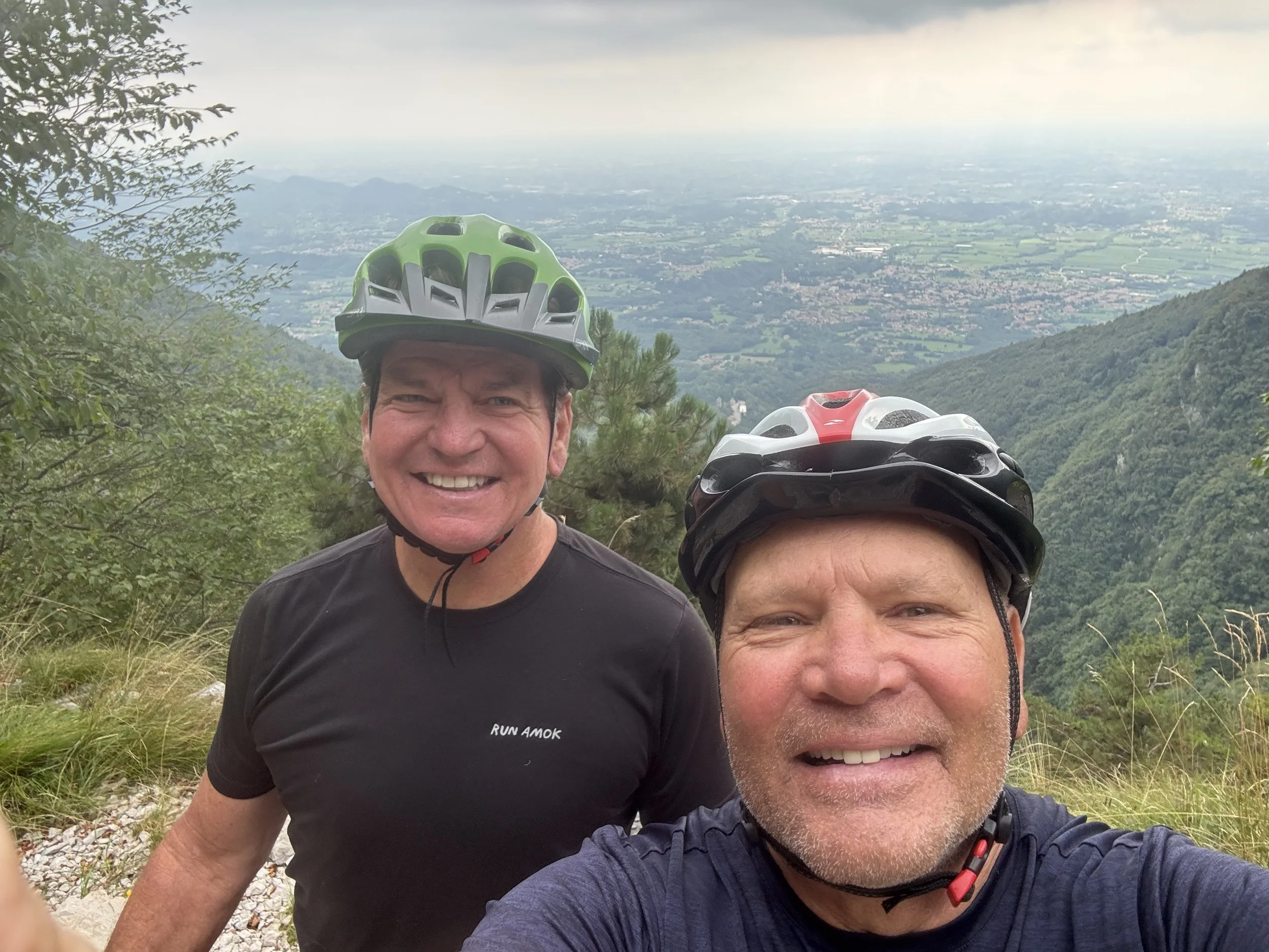 Two men taking a selfie on a mountain trail with a scenic view of the valley and hills in the background. They are wearing bicycle helmets and athletic clothing.