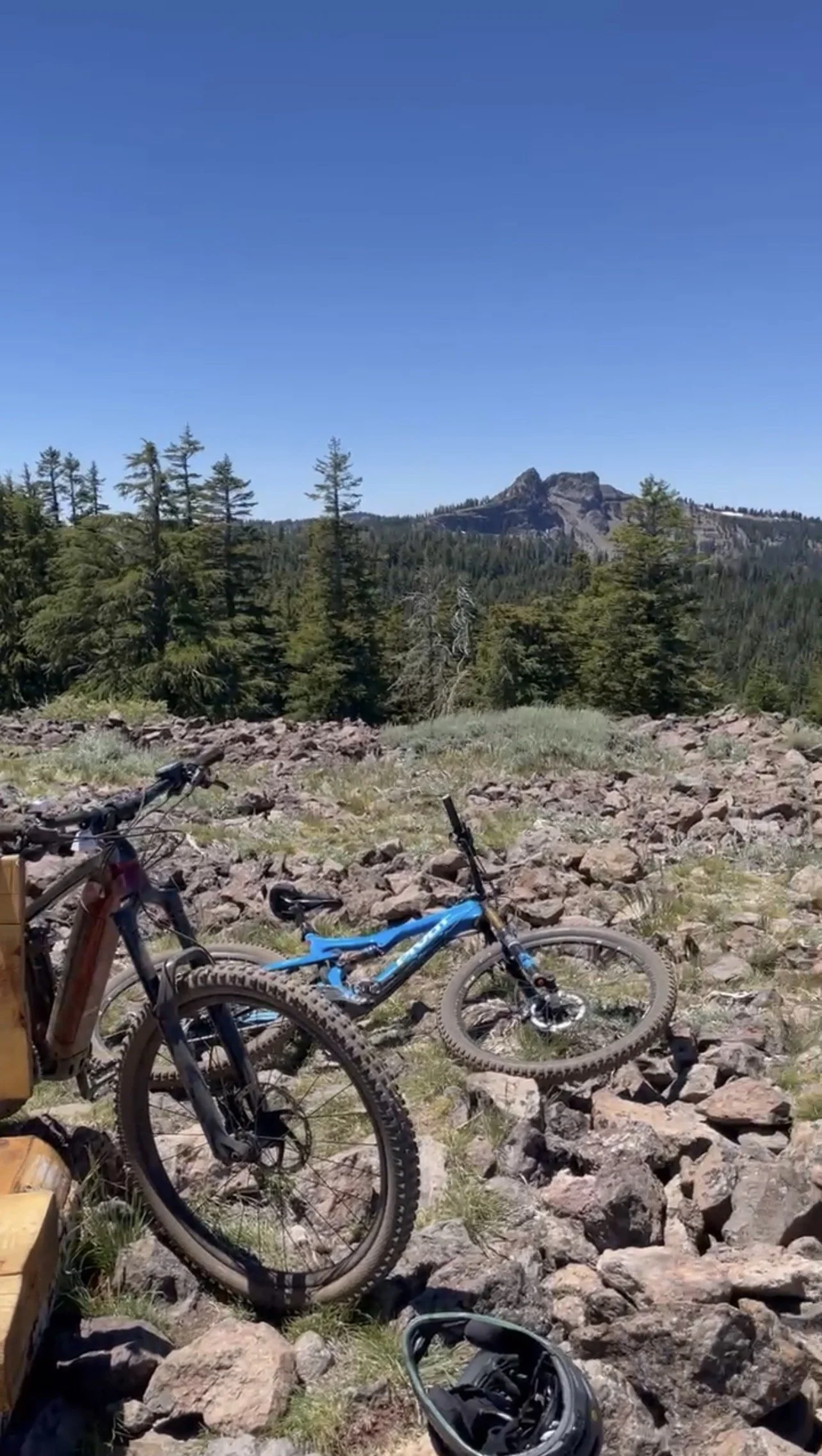 Mountain bikes resting on rocky ground with a forest and mountain in the background under a clear blue sky.