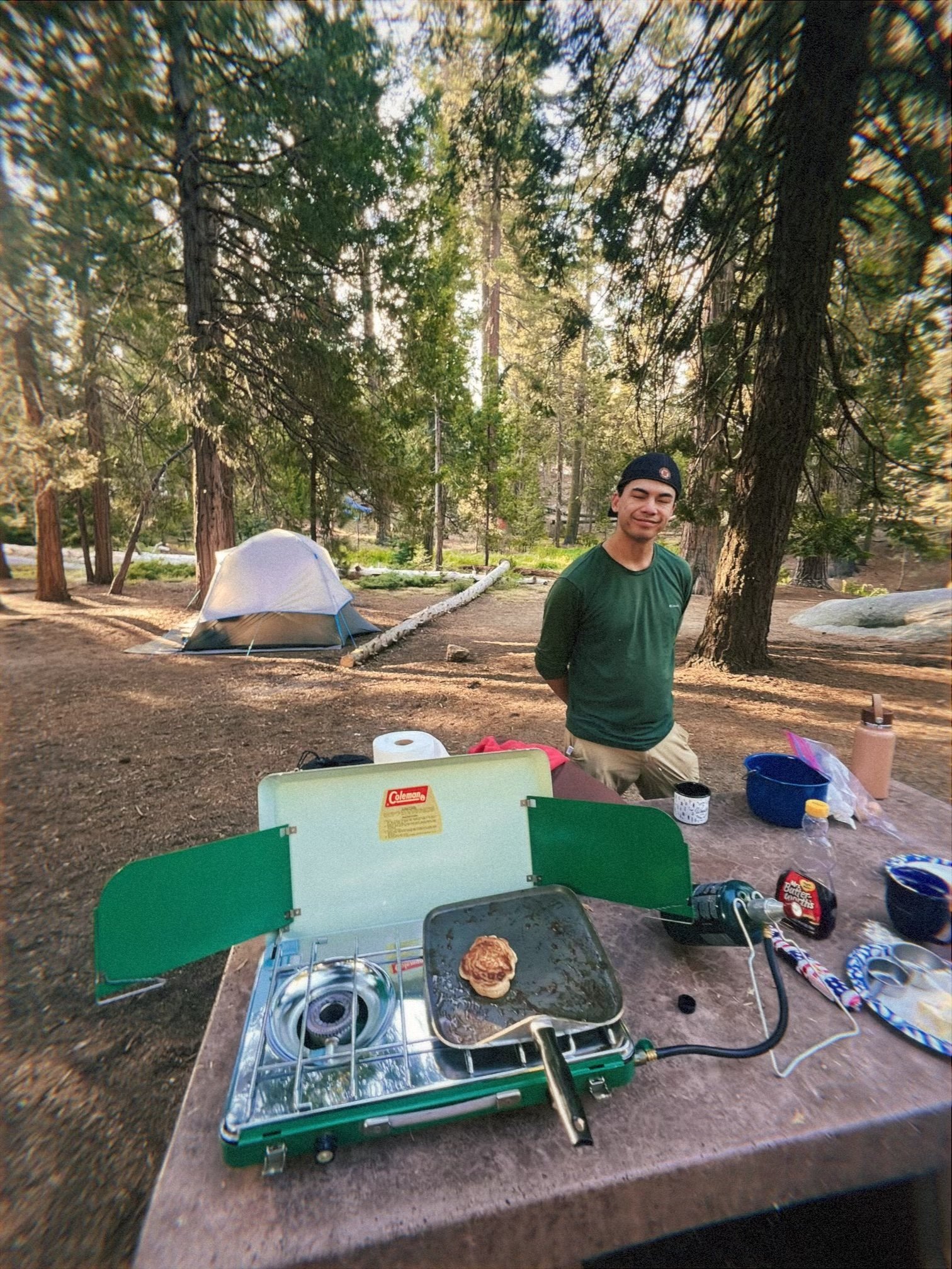A man standing outdoors in a forested area with camping tents in the background. There is a portable stove with a bun on it, and camping supplies on a wooden table in the foreground.