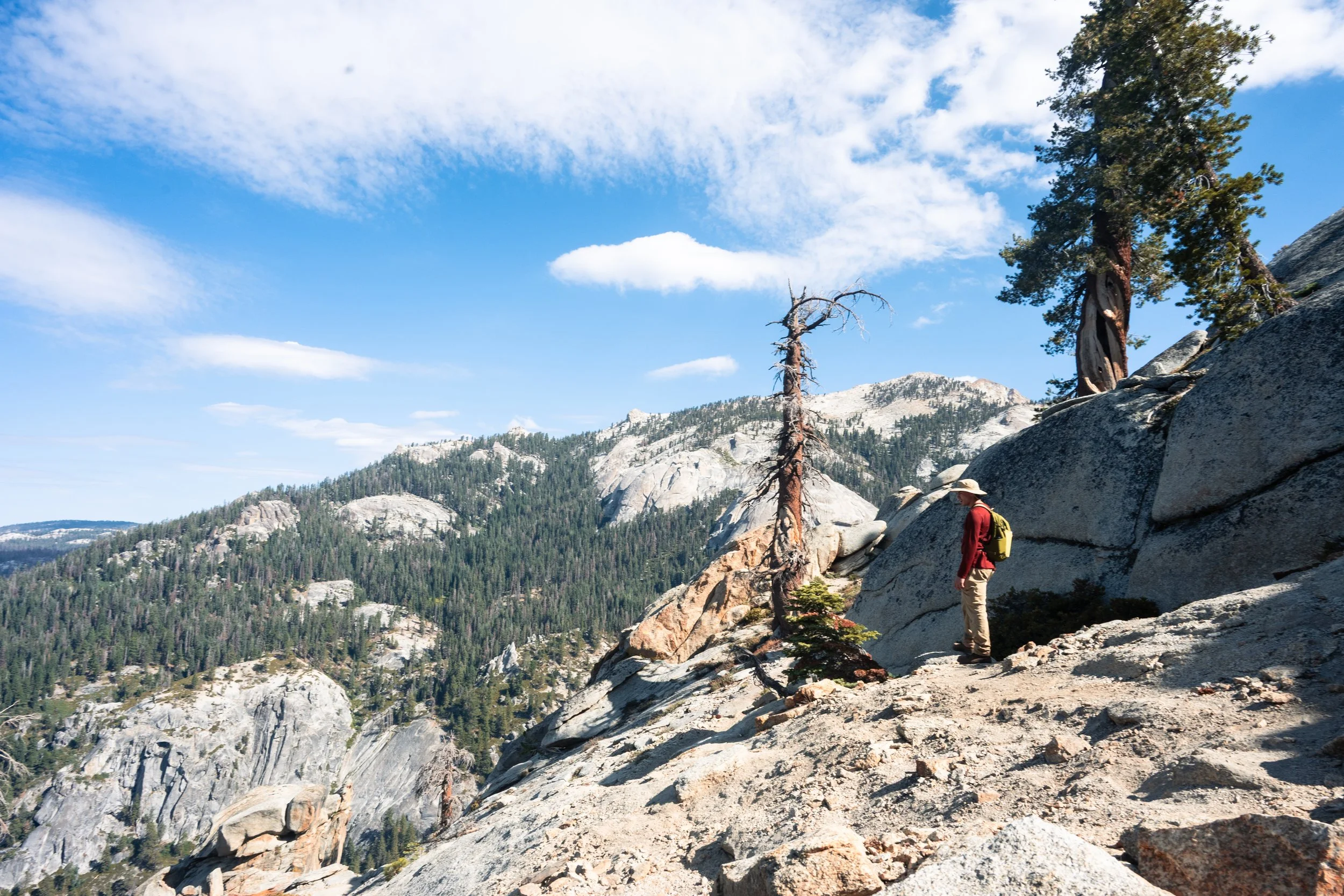 A hiker with a yellow backpack and hat walking along a rocky mountain trail. The background features forested mountains, evergreen trees, and a partly cloudy blue sky.