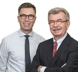 Zwei Herren in formeller Kleidung, einer älter, der andere jünger, vor weißem Hintergrund. Beide Optiker und Kunden des OptikamPad Zentriersystems