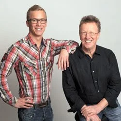 Zwei lachende Männer, einer jüngerer mit Brille und Hemd, der andere älter mit dunklem Hemd, posieren vor grauem Hintergrund. Beide sind Optiker und Kunden des OptikamPad Zentriersystems