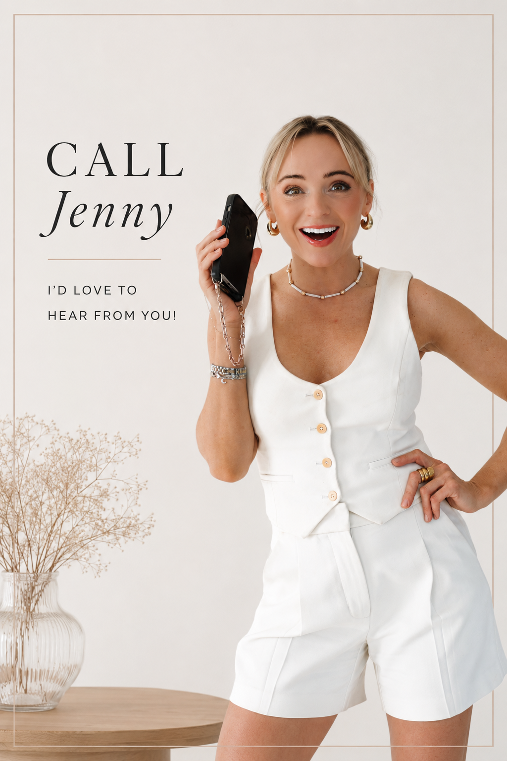 A woman in a white sleeveless outfit holding a black phone with a chain, smiling, with a white background and a vase with dried flowers on a table.