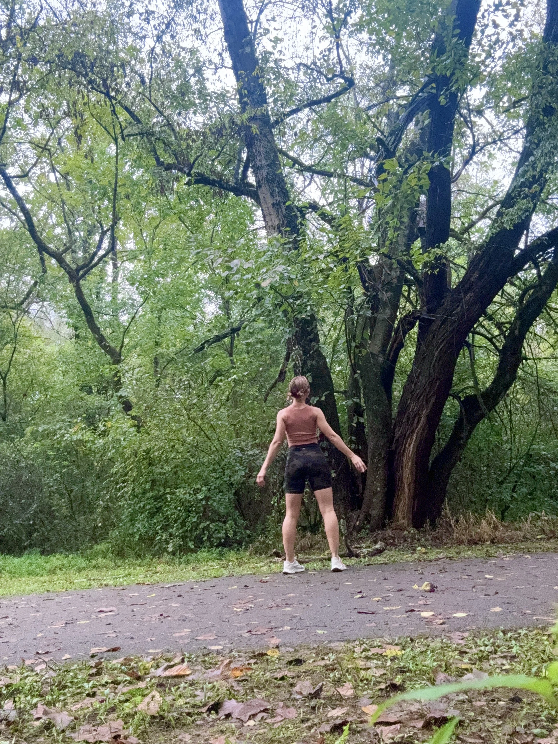 A person with short hair wearing a sleeveless top, shorts, and white shoes stands facing away from the camera in a wooded park area, with trees and foliage surrounding them.