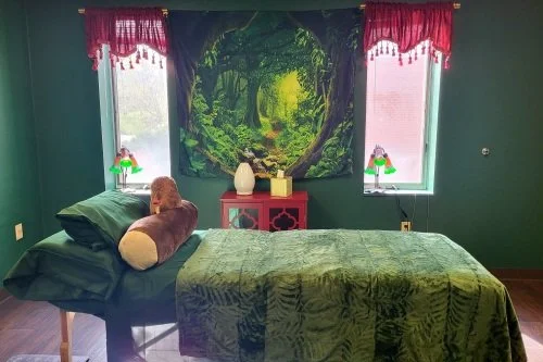 A clinical space with a massage table covered in green blankets and pillows and a nature-themed tapestry on the wall behind it. There are two windows with red curtains and flower shaped lamps. A red table in the middle has a glass diffuser.