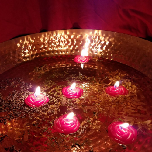 Five floating red rose candles in a copper bowl filled with water and herbs, reflecting warm light.