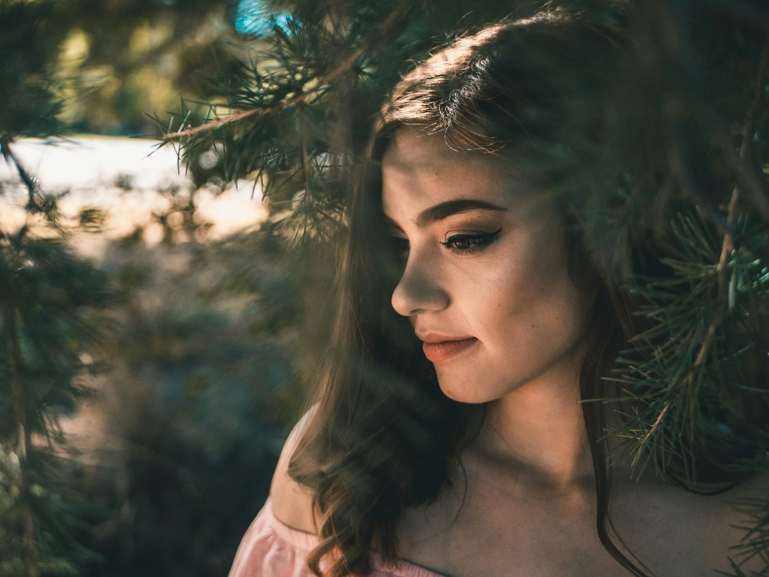 Close-up of a woman's face with long wavy hair and makeup, surrounded by pine tree branches in a natural outdoor setting.
