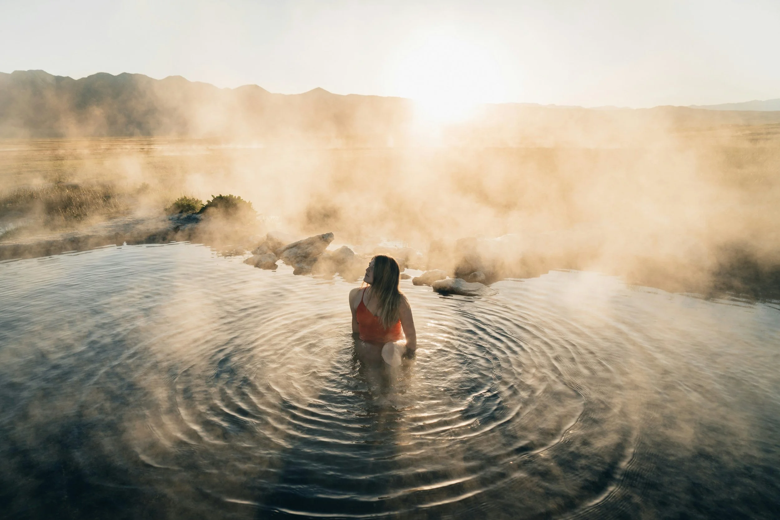 A woman in a red swimsuit soaking in hot spring water with steam rising and mountains in the background at sunrise or sunset.