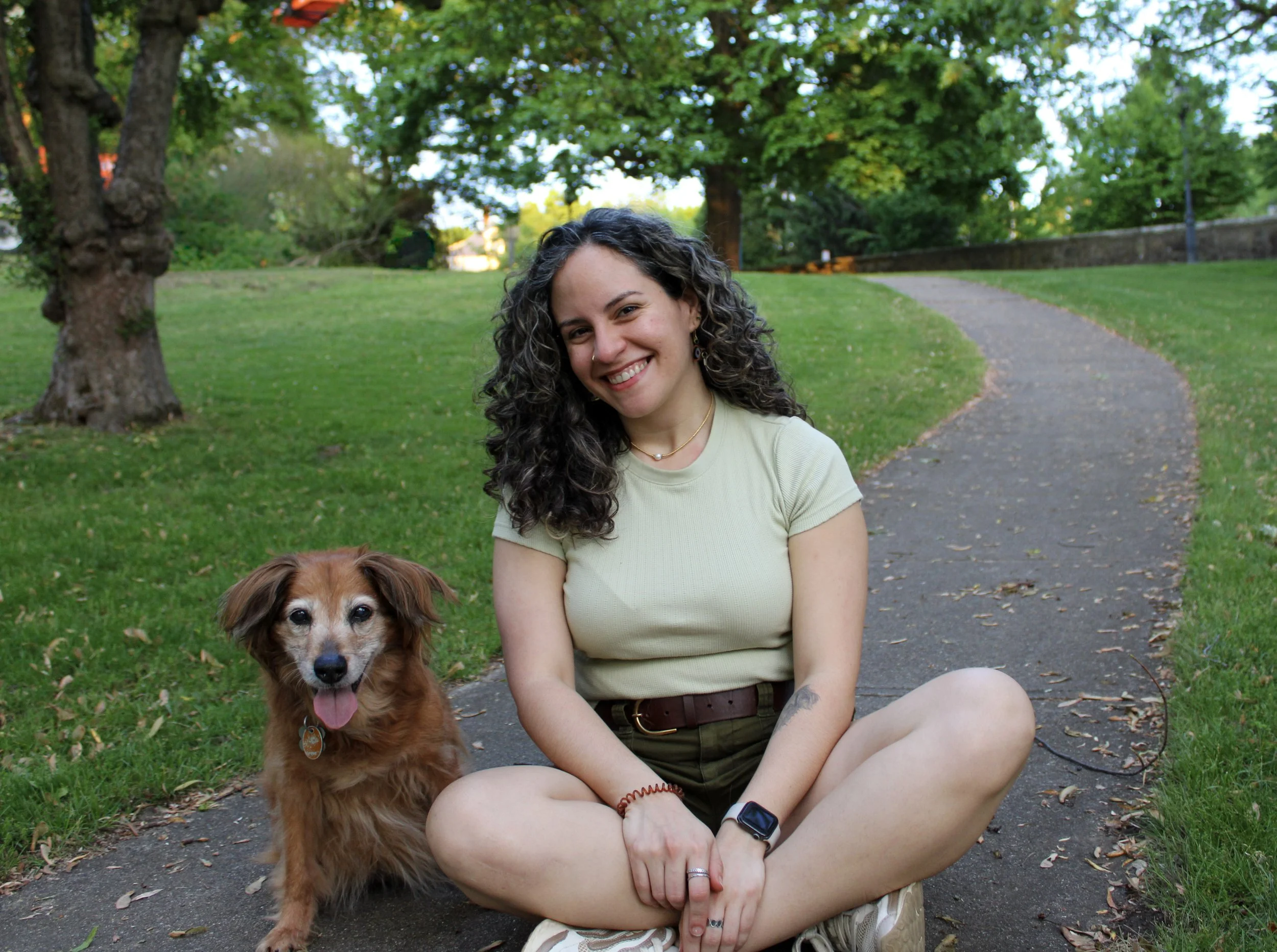 A pet sitter woman smiling while sitting cross-legged on a walking path in a park, accompanied by a brown dog.