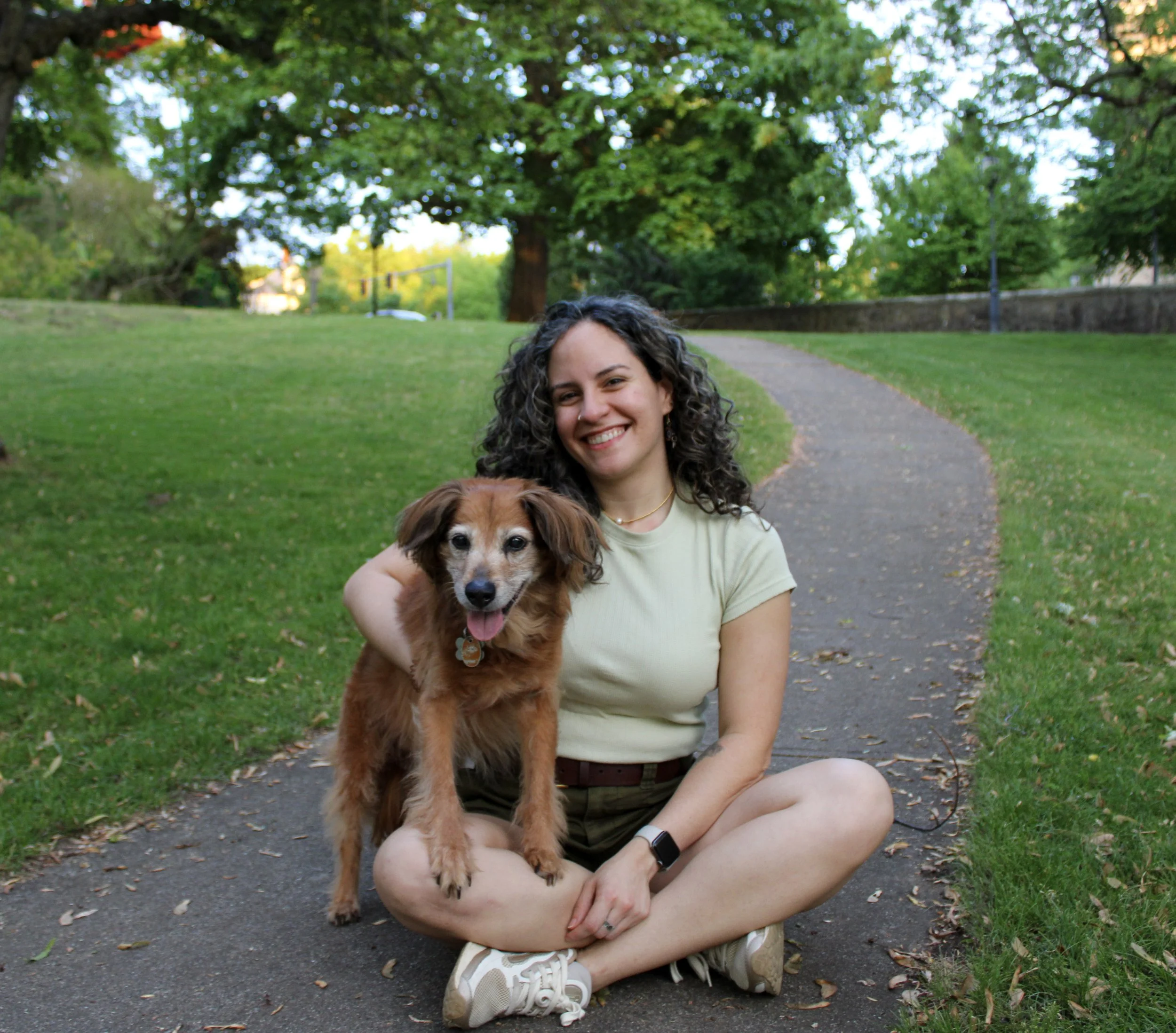 A woman pet sitter sitting on the sidewalk in a park, holding a brown dog. There are green trees and a grassy area in the background.